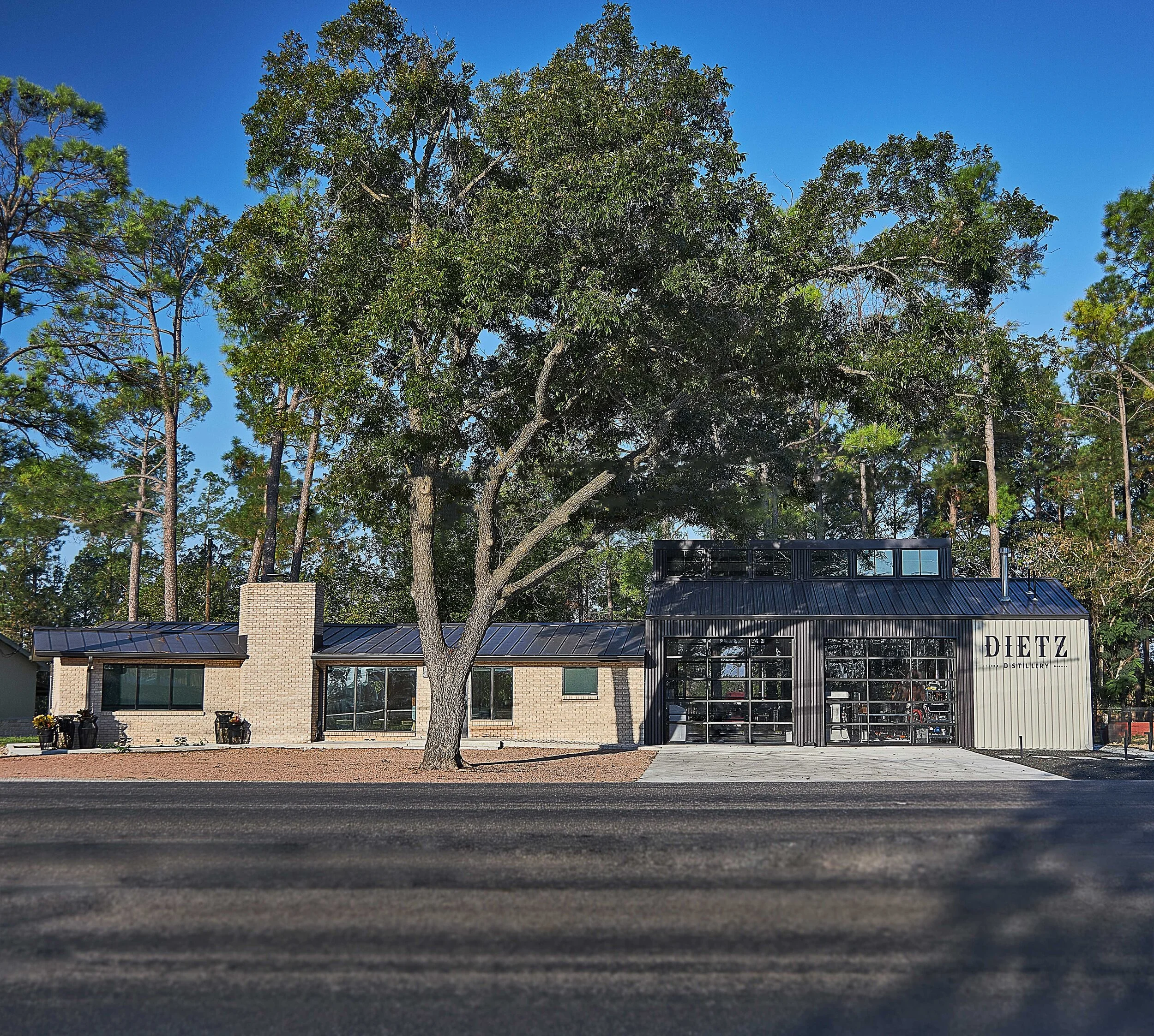 A modern building with large glass windows and a black metal roof, in front of tall trees with green foliage under a clear blue sky.
