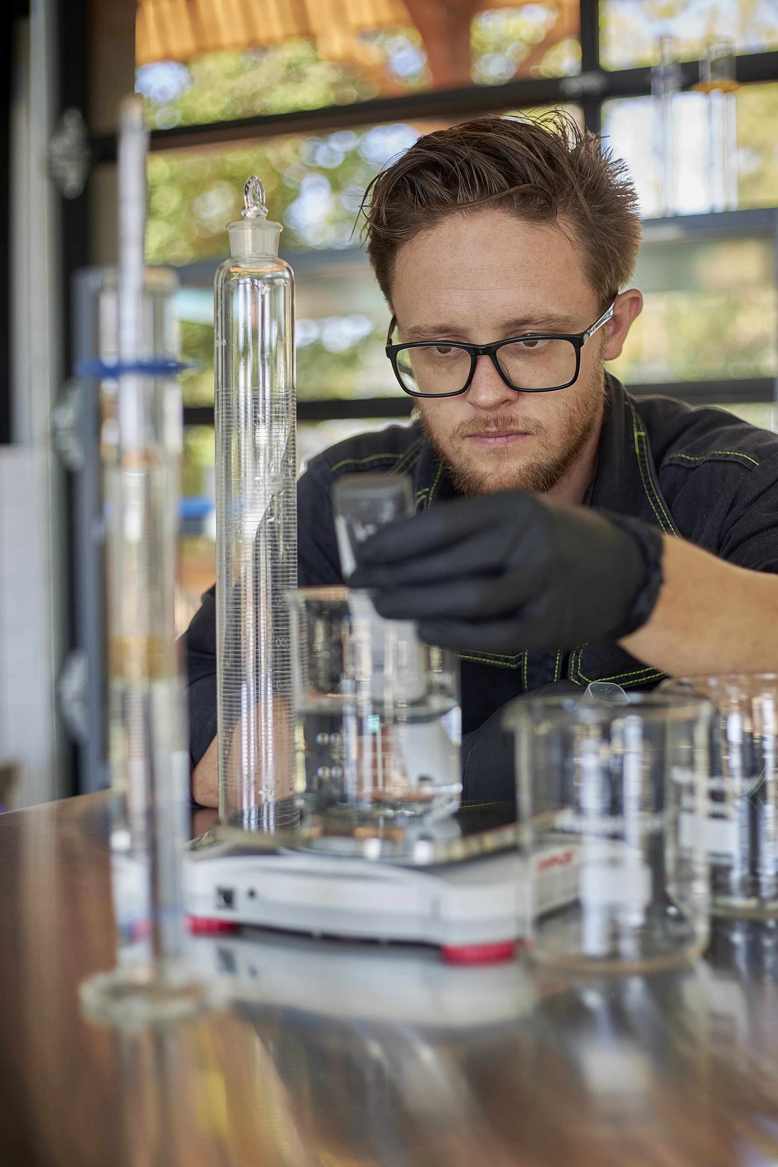 A man with glasses and a beard working with laboratory glassware, including graduated cylinders, in a well-lit lab or workshop with large windows showing trees outside.