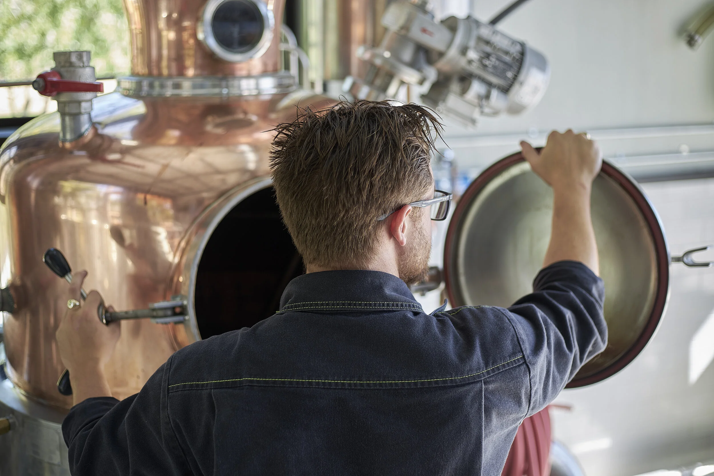 A man with glasses and brown hair, wearing a dark jacket, is turning a large copper-colored metal valve or lid.