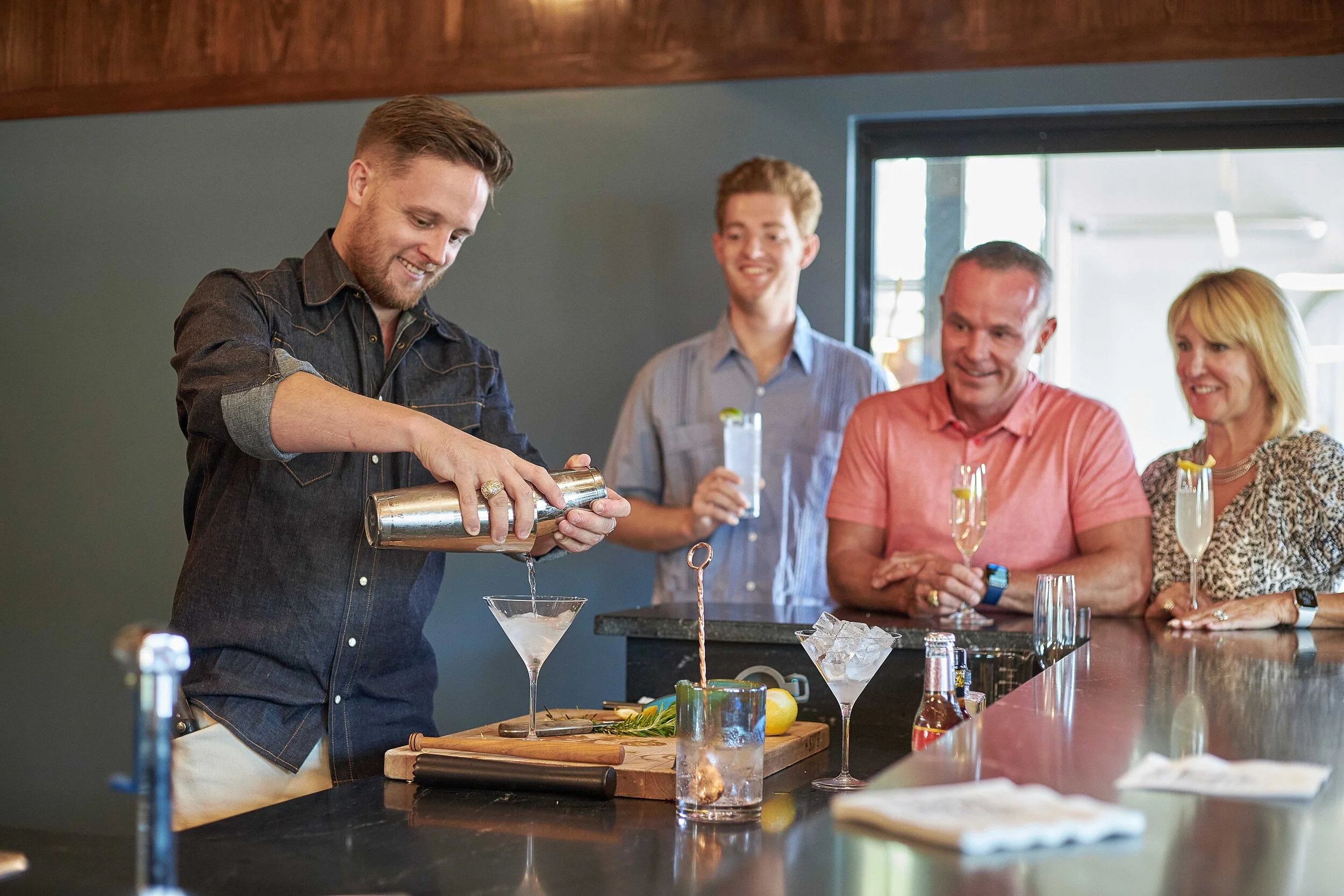 A man creates cocktails at a bar while four people watch and smile, holding drinks. The bar has various bottles and garnishes, and the setting appears lively and social.