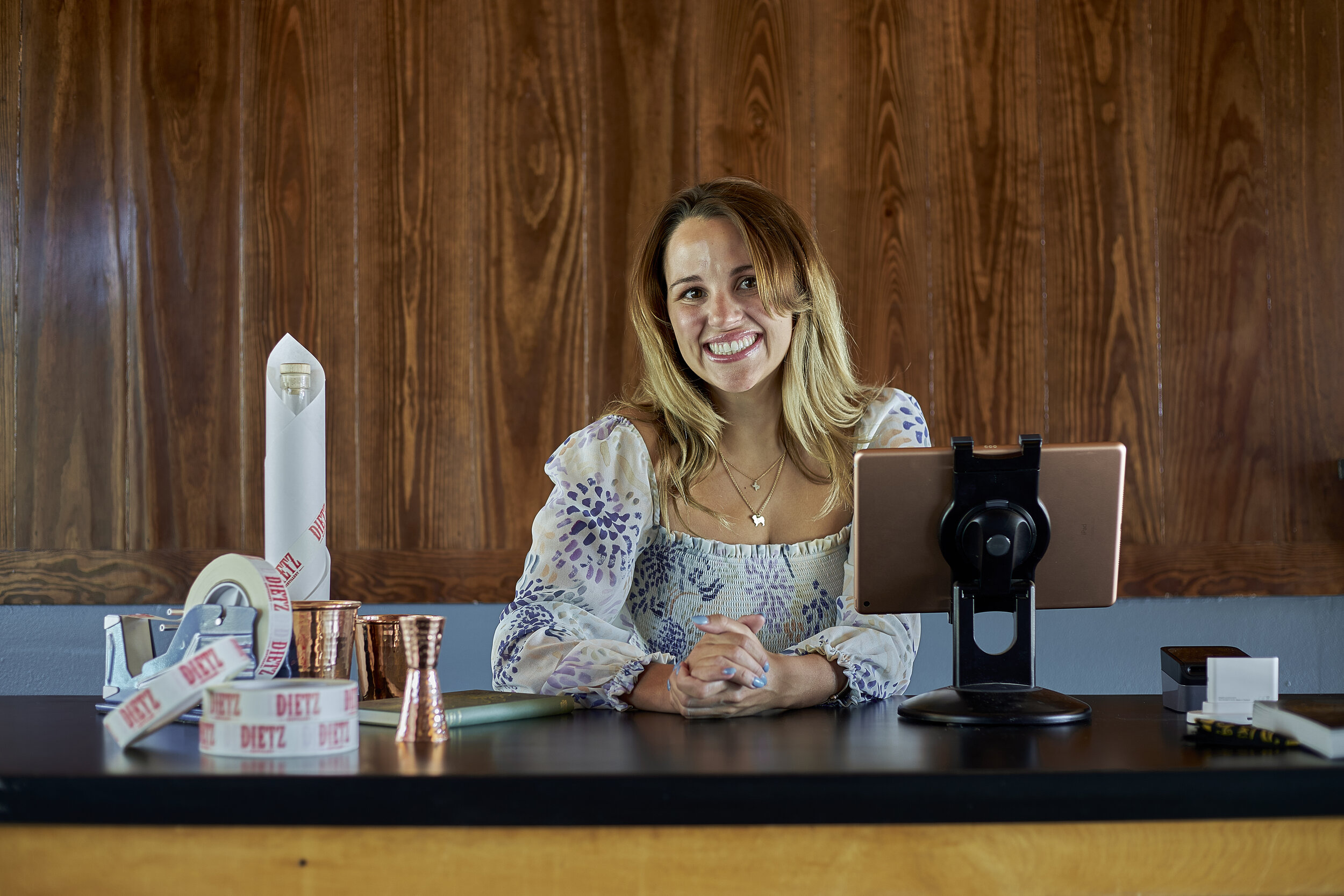 Woman with blonde hair smiling, sitting at a table with drink cups, tape, and a tablet on a stand, in front of wooden paneling.