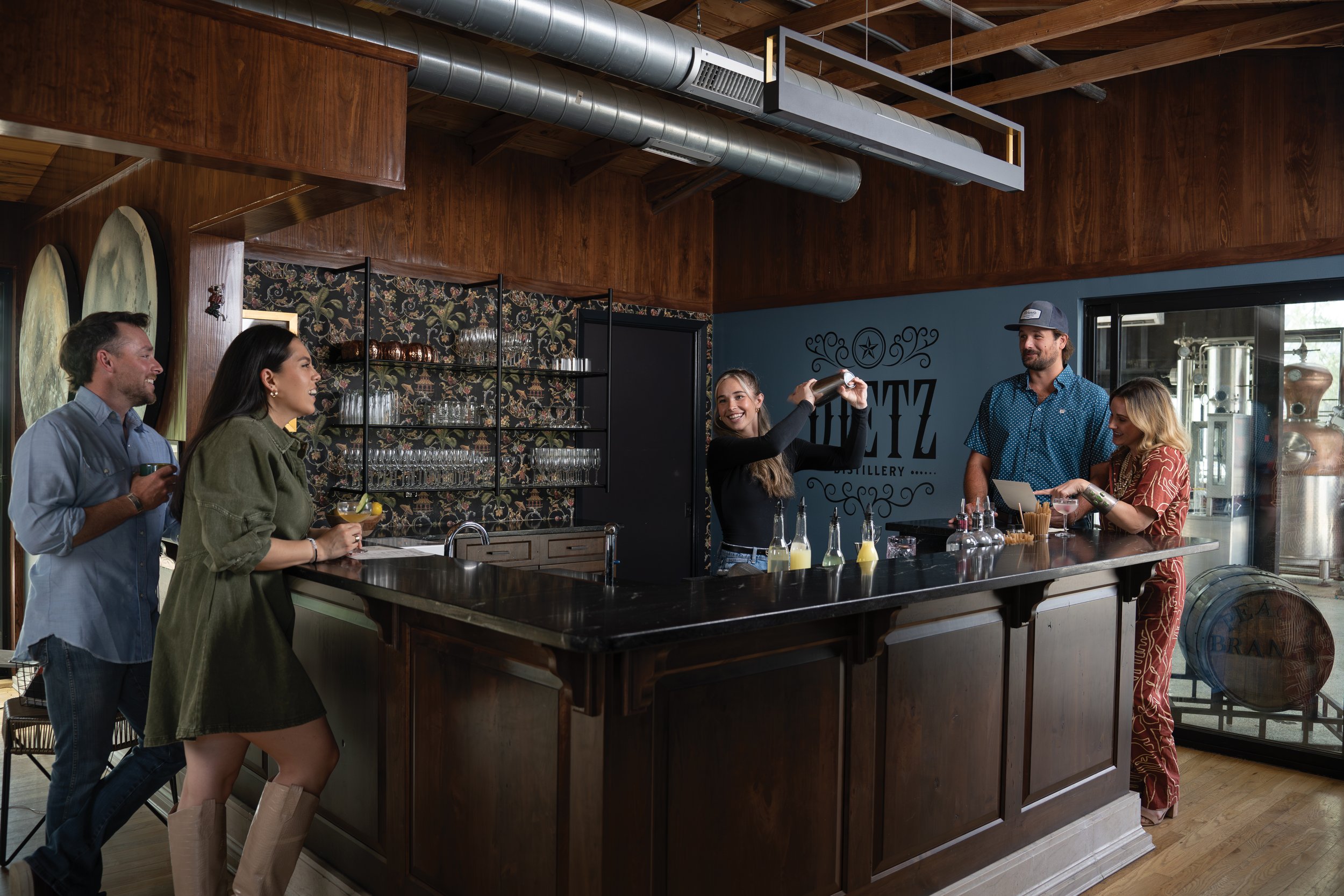 A group of people enjoying drinks and conversation at a bar inside a brewery. A woman is bartending, preparing drinks behind the counter, while four customers stand nearby, smiling and engaging with the bartender.