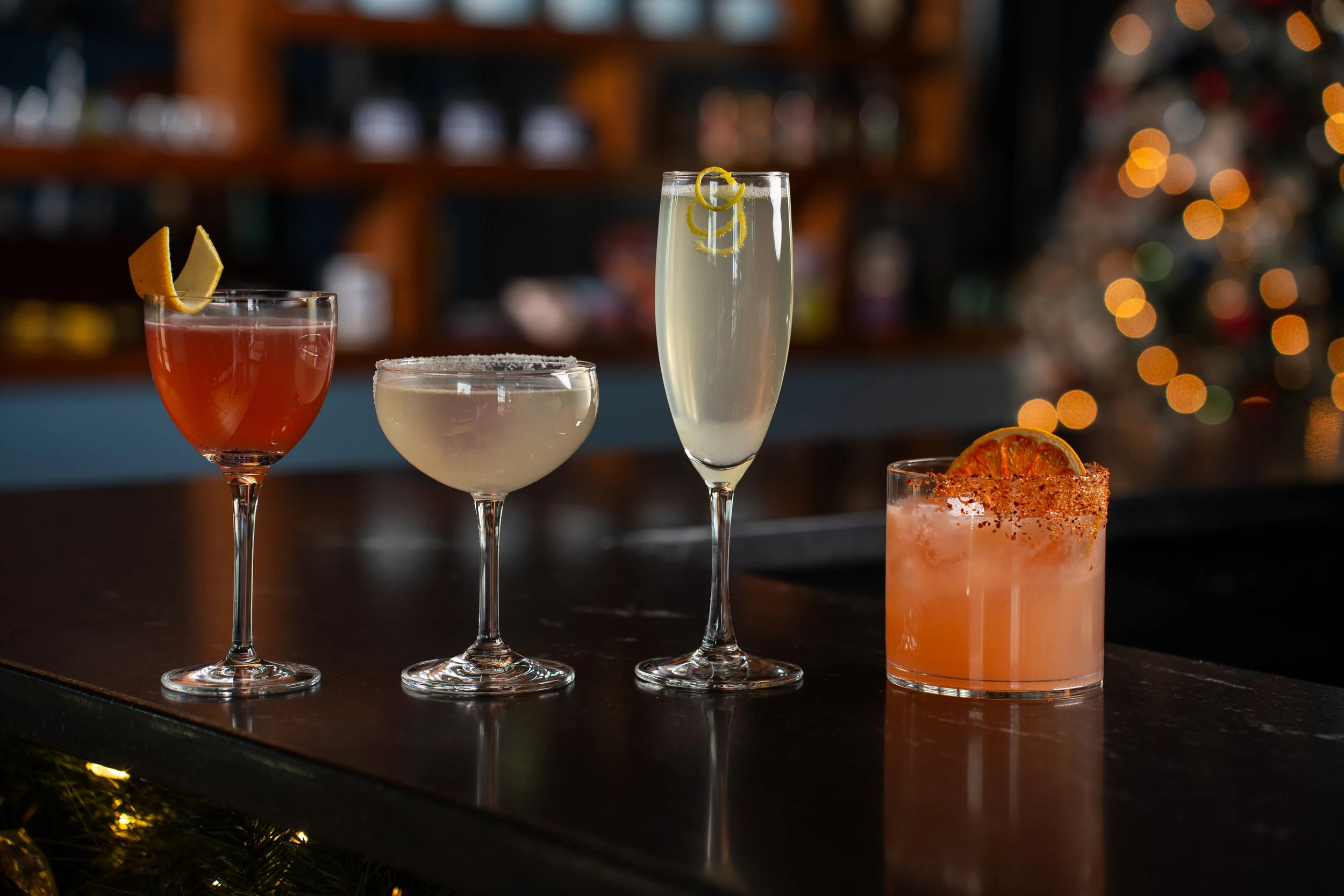 Various cocktails displayed on a bar counter with a blurred background of bartending area and festive orange bokeh lights.