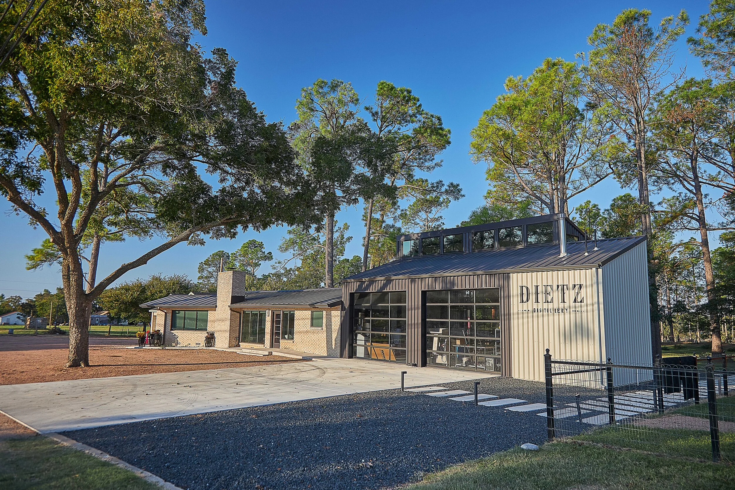 A modern distillery building with large windows and a sign that reads 'DIETZ Distillery,' surrounded by tall trees and a gravel yard, under a clear blue sky.