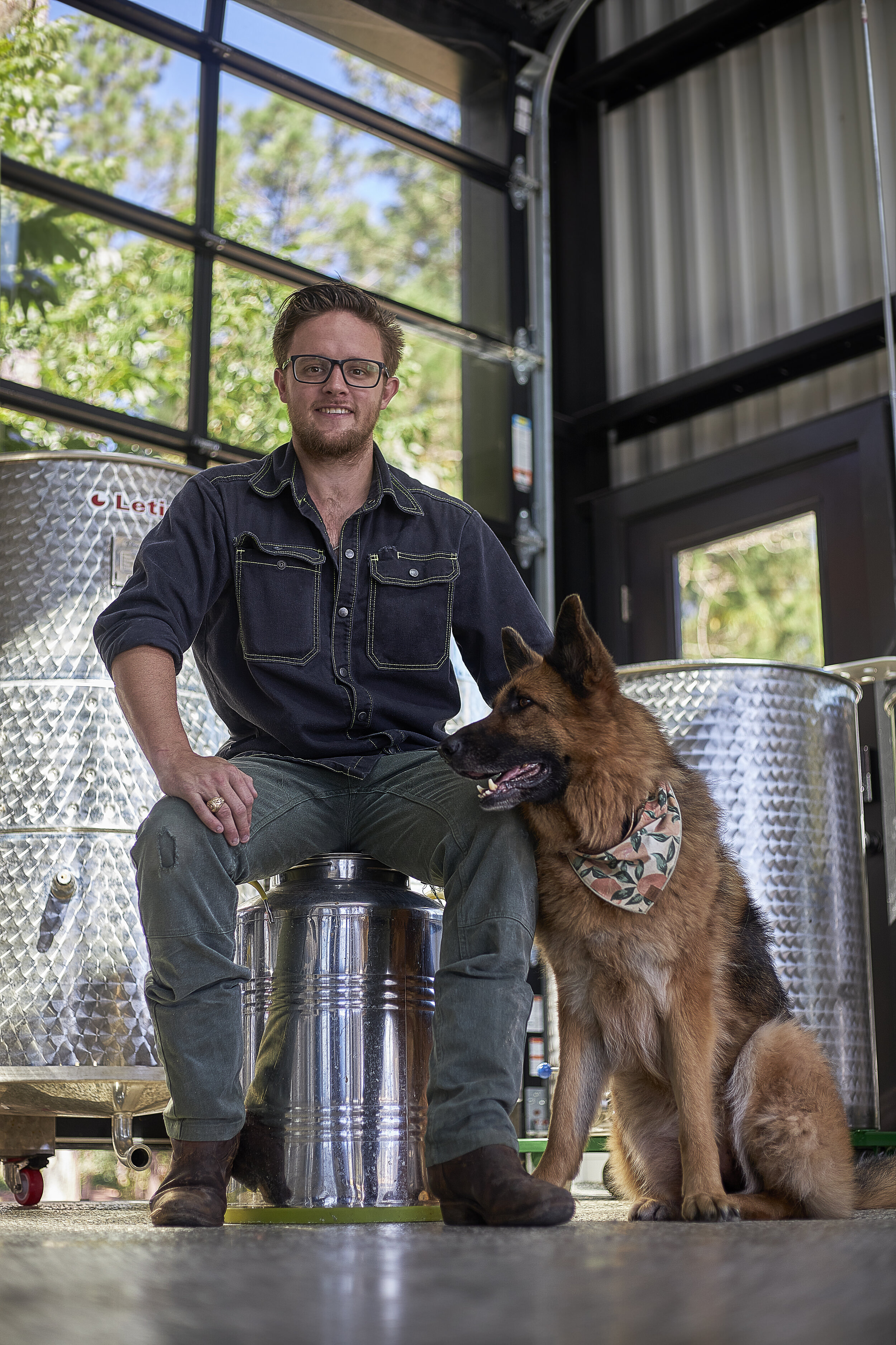 A man with glasses and a beard, wearing a dark denim shirt and jeans, sitting in a workshop with his dog. The dog is a large German Shepherd sitting beside him, wearing a bandana. The workshop has metal tanks and large windows with greenery outside.