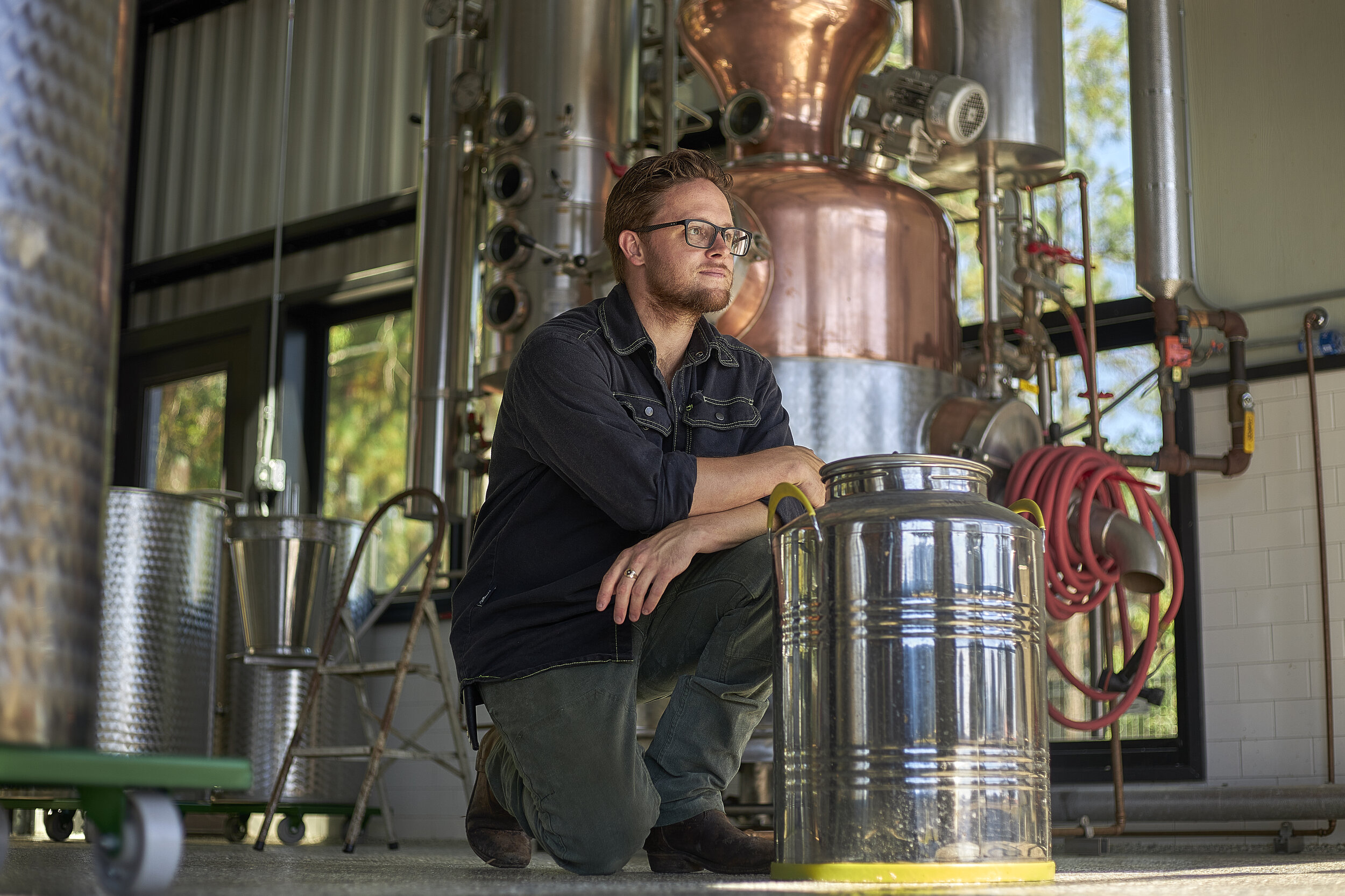 A man with glasses and a beard sitting on the floor in front of an industrial distillation setup, looking thoughtfully to the side. The setup includes a copper still and various pipes and gauges. Sunlight illuminates the area through large windows, a