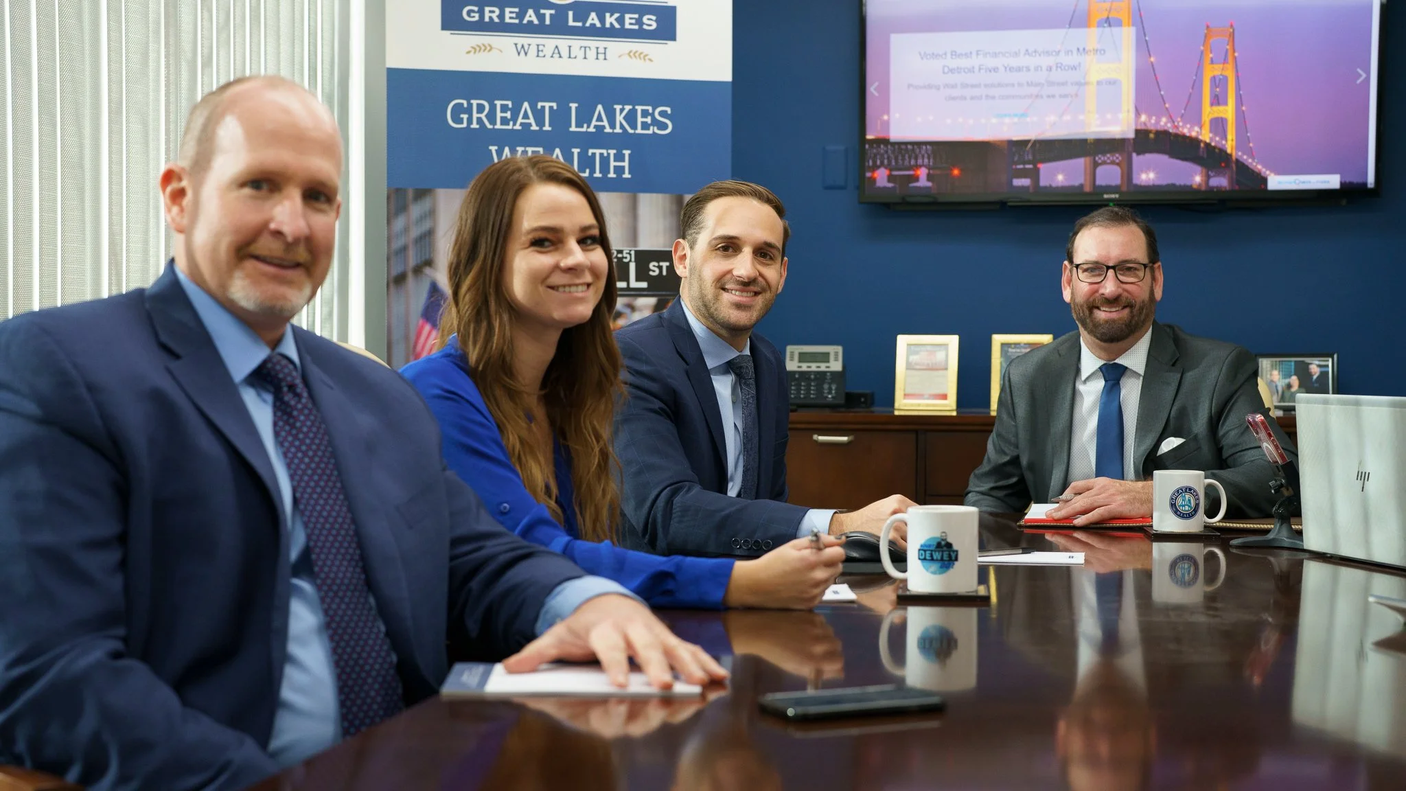Four professionals sitting at a conference table, smiling, with a blue and white promotional backdrop reading 'Great Lakes Wealth' and a TV screen showing a bridge. They have coffee mugs and notebooks in front of them.