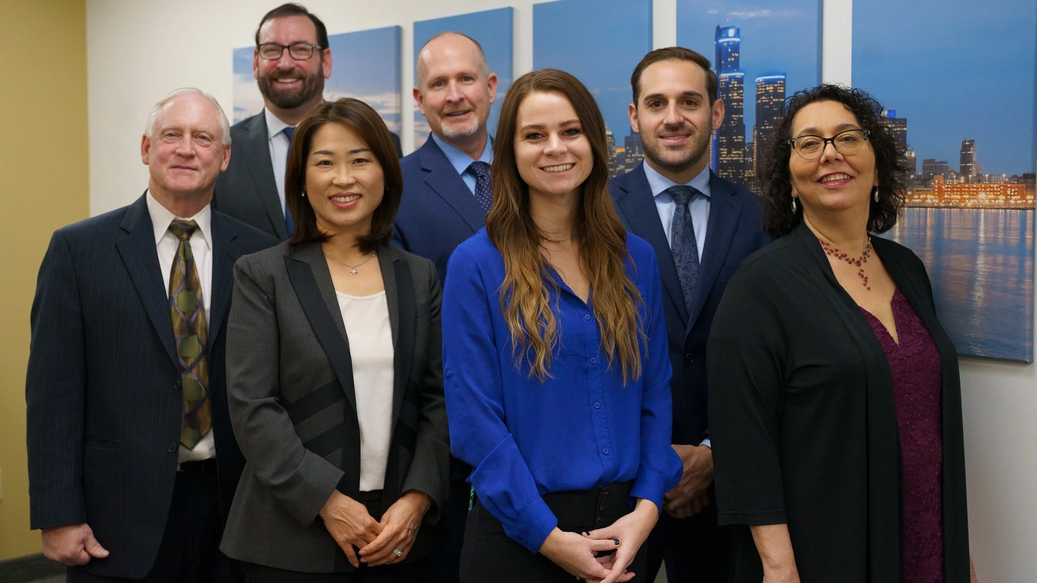 Group of seven professionally dressed people posing in an office with cityscape artwork in the background.
