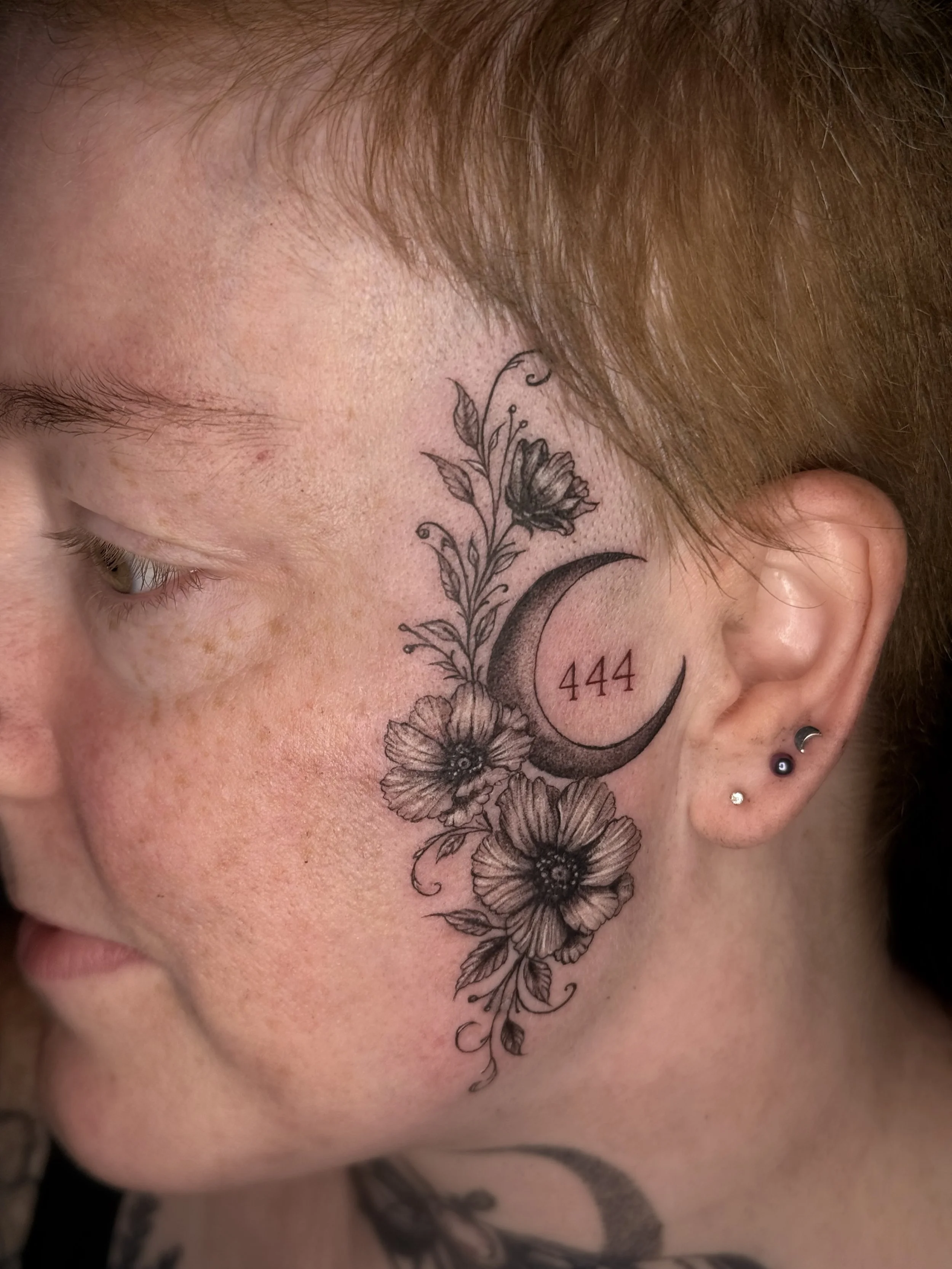 Close-up of a person's face with tattoos on the side of their face, including a crescent moon, flowers, and the numbers 444, with reddish hair and earrings.