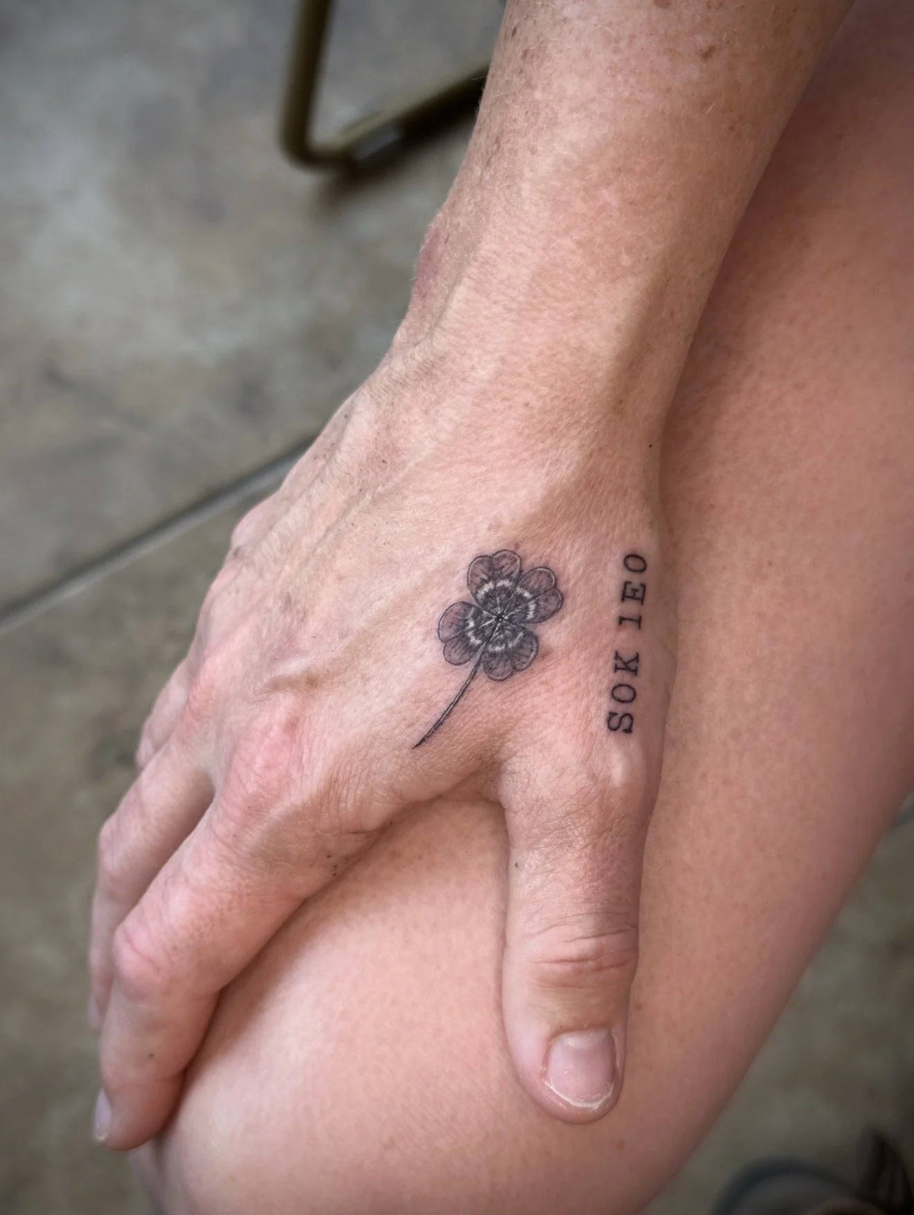 Close-up of two hands softly clasped on a person's lap, showing a tattoo of a four-leaf clover and the text 'SOX 10' on the wrist.