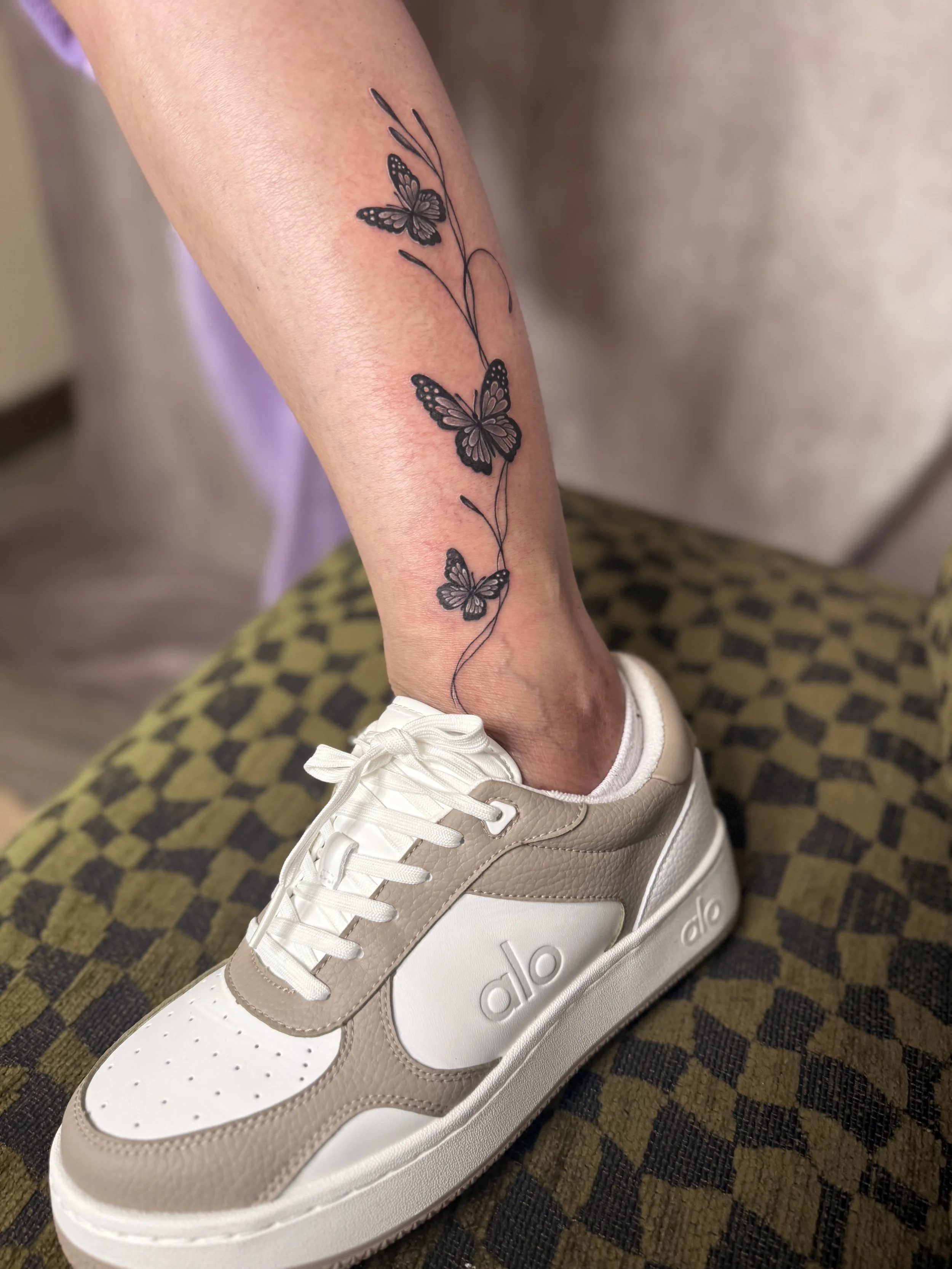 Tattoo of three butterflies connected by a line on a person's forearm, with part of a beige sneaker visible.