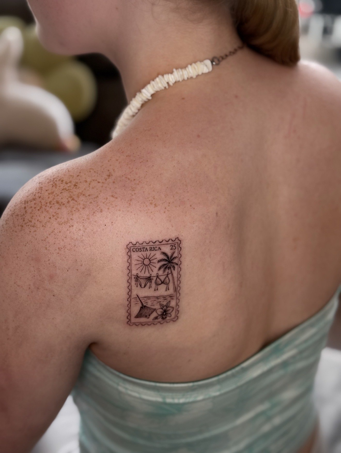 A person with fair skin and reddish hair, wearing a shell necklace, has a tattoo of a Costa Rican postage stamp on their shoulder. The tattoo depicts a tropical scene with a sun, palm trees, a bikini-clad woman hanging laundry, and a flower.