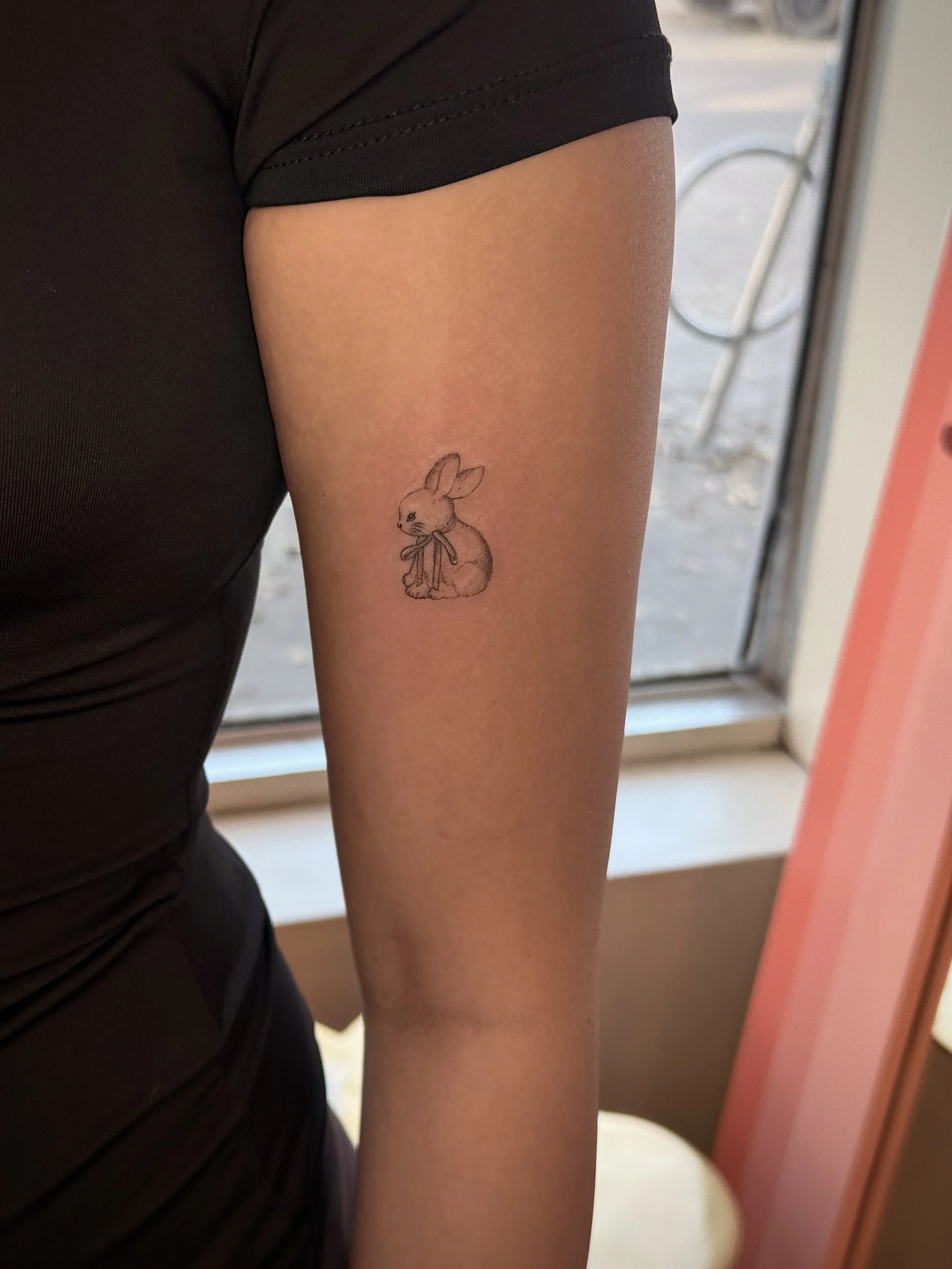 A person's upper arm showing a small bunny tattoo with a bow, next to a window.