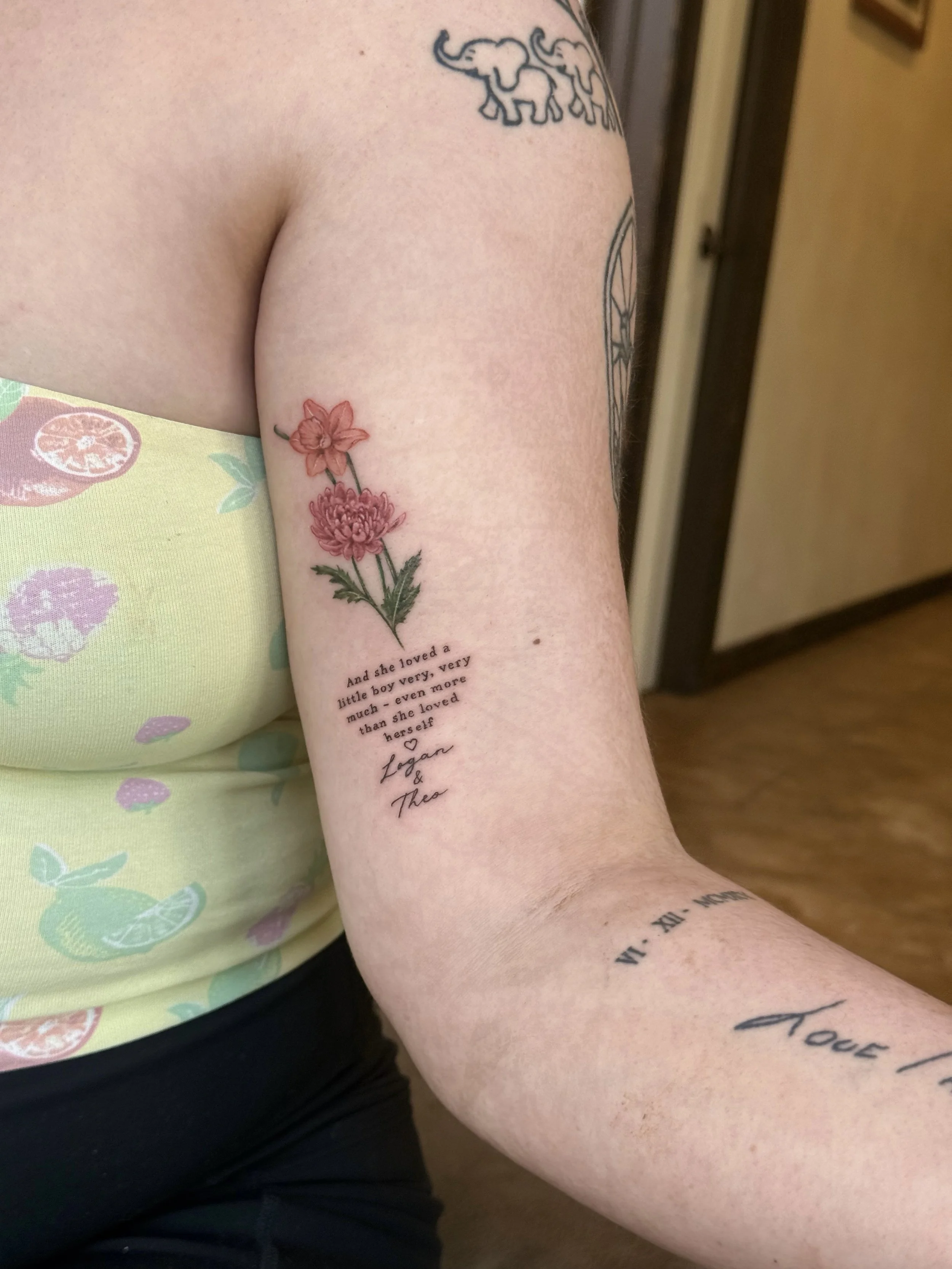 Close-up of a person's arm with multiple tattoos, including a floral tattoo of pink flowers, a quote, and other designs, worn while sitting on a floral-patterned surface, with a background of a wooden floor and cabinet.