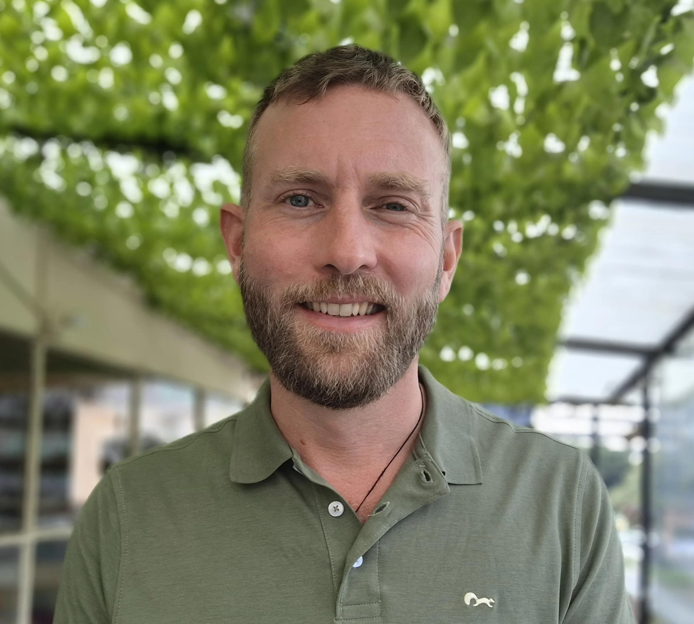 A photograph of a handsome blonde man with a tidy beard standing outside. The man is wearing a green polo shirt.