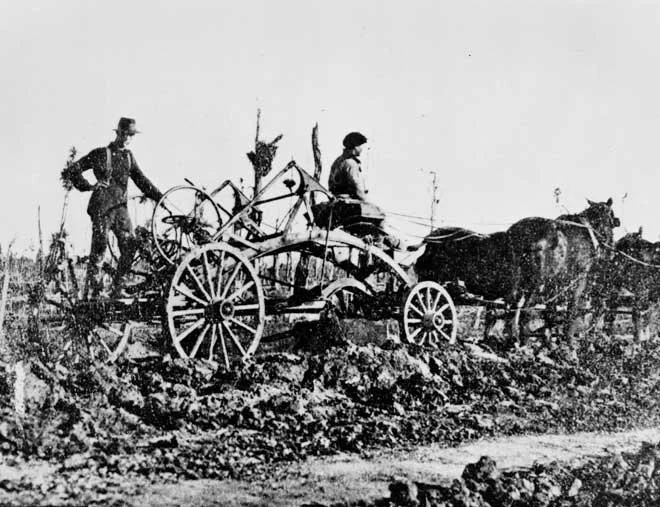An old black-and-white photograph of a horse pulling a piece of machinery in a muddy, rough environment. Two men stand aboard the machine, one with reigns to the horse and the other facing the camera.