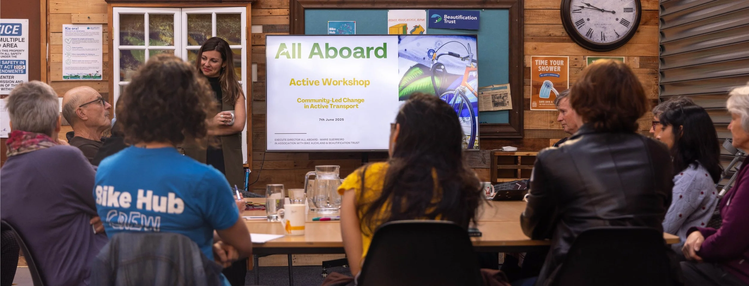 A group on people seated at a table. In front of them, a young woman presents on a large screen. The screen reads "All Aboard Active Workshop"