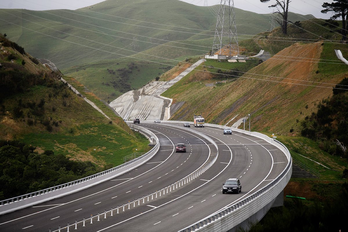 A photograph of a large motorway cutting through green hills. The road has wide lanes, wide shoulders, and barriers on both sides as well as in the centre of the road. There is very little traffic on a very large road.