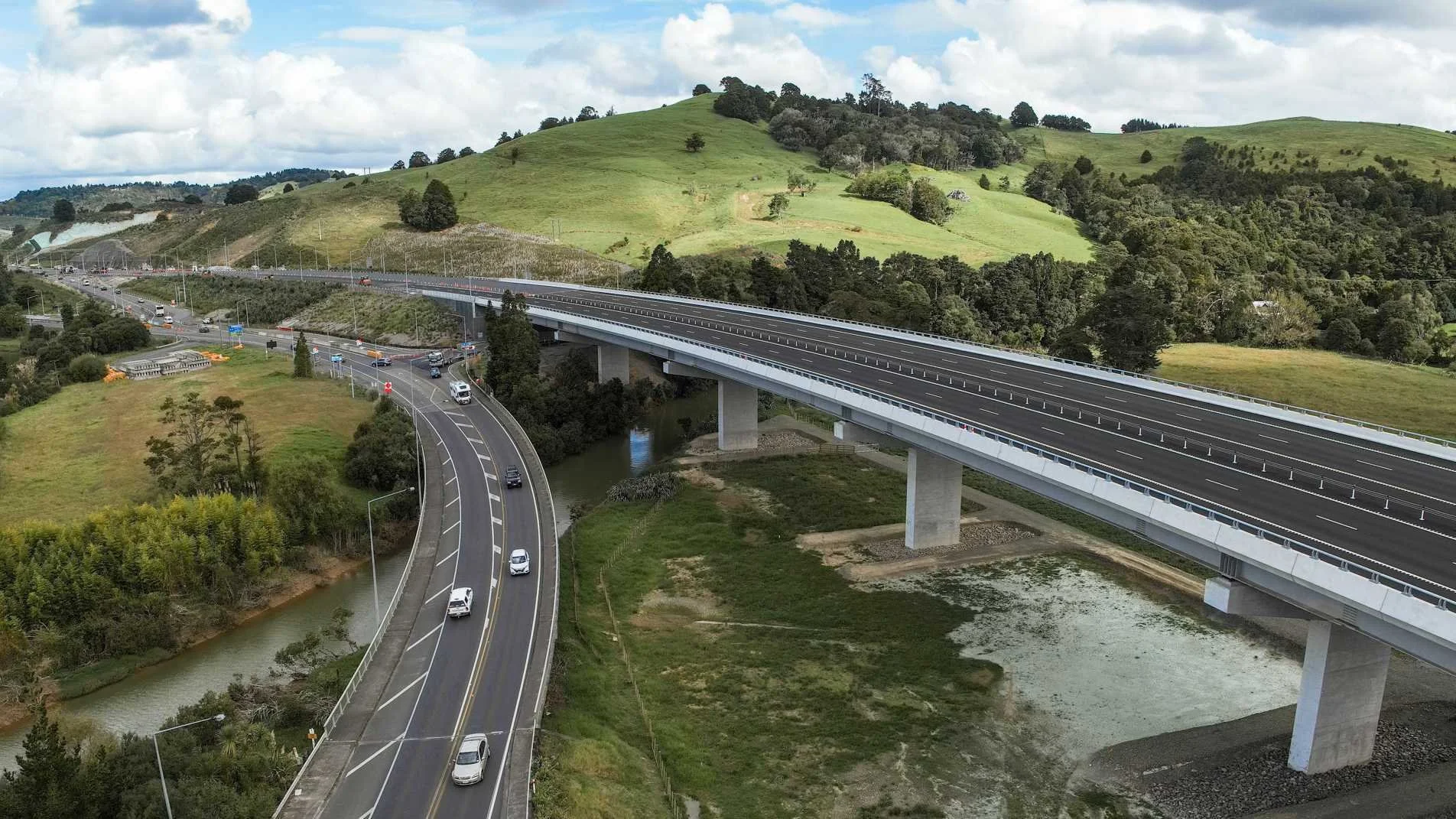 A photograph showing two roads. One smaller road is at a lower level, curving as it crosses a river. A much larger, newer road is elevated above both the smaller road and the river.