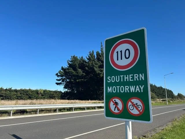 A photograph of a 110 kilometre per hour speed sign against a backdrop of a major road and blue sky.