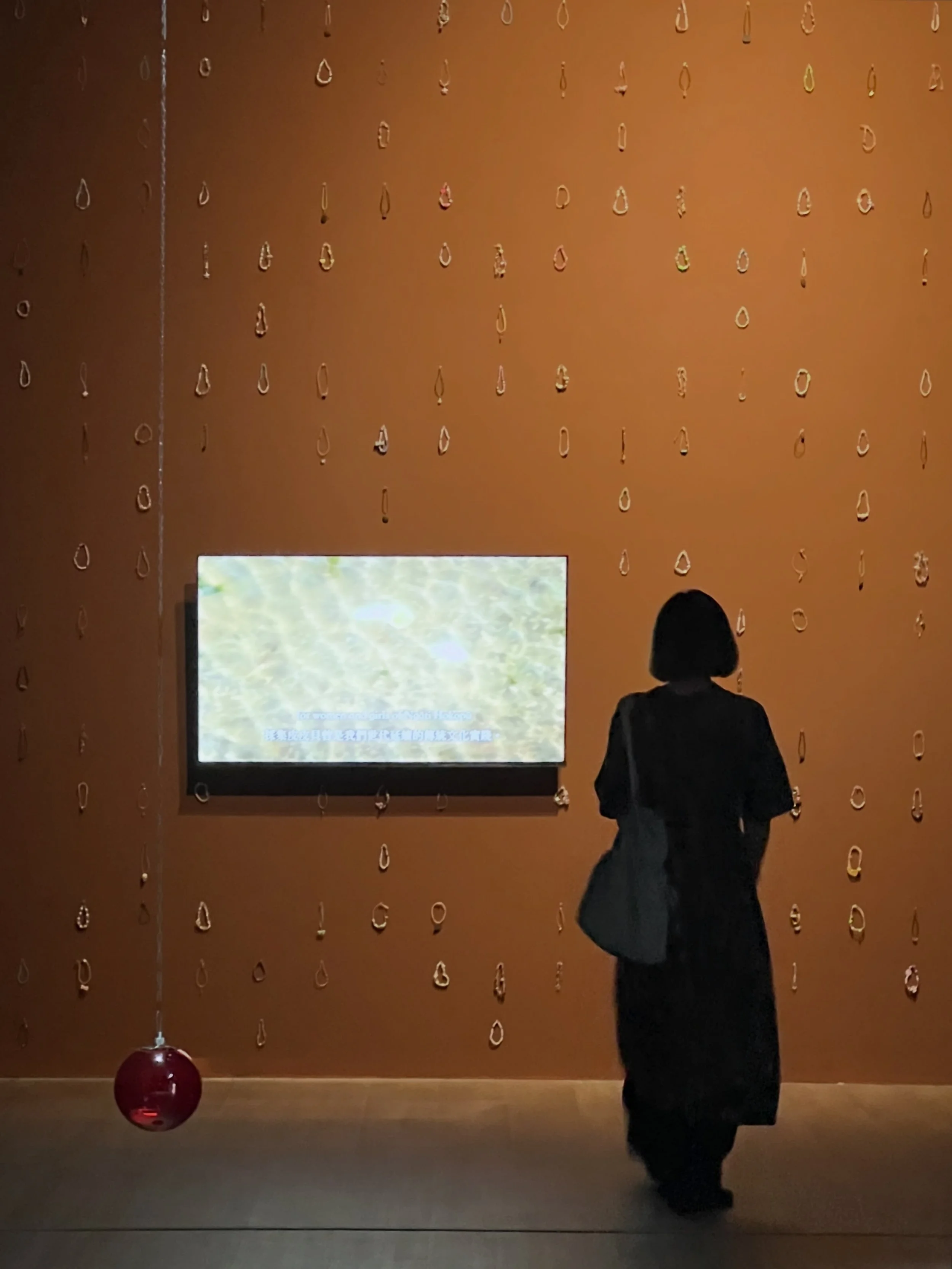 Installation View of Middens made by she/shells. Friendship bracelets attached to the wall in a pattern imitating shells. A video screen on a brown wall.