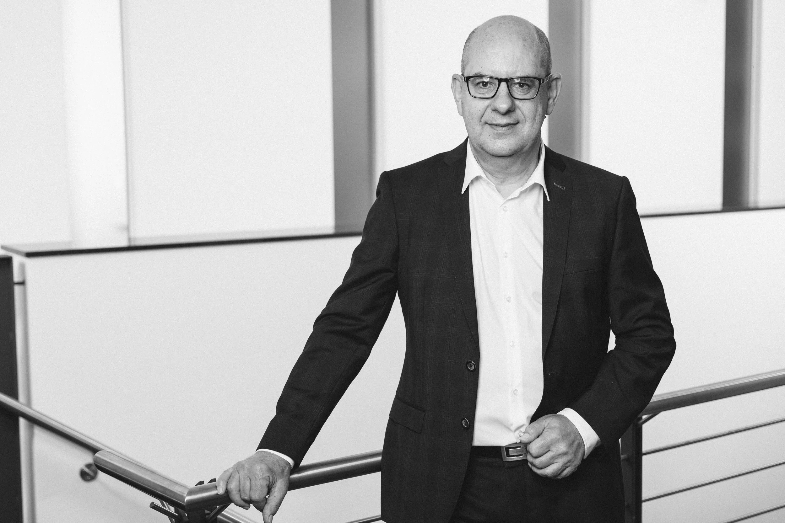 Mann in dunklem Anzug mit Brille lehnt an einem Geländer in einem modernen Büro.