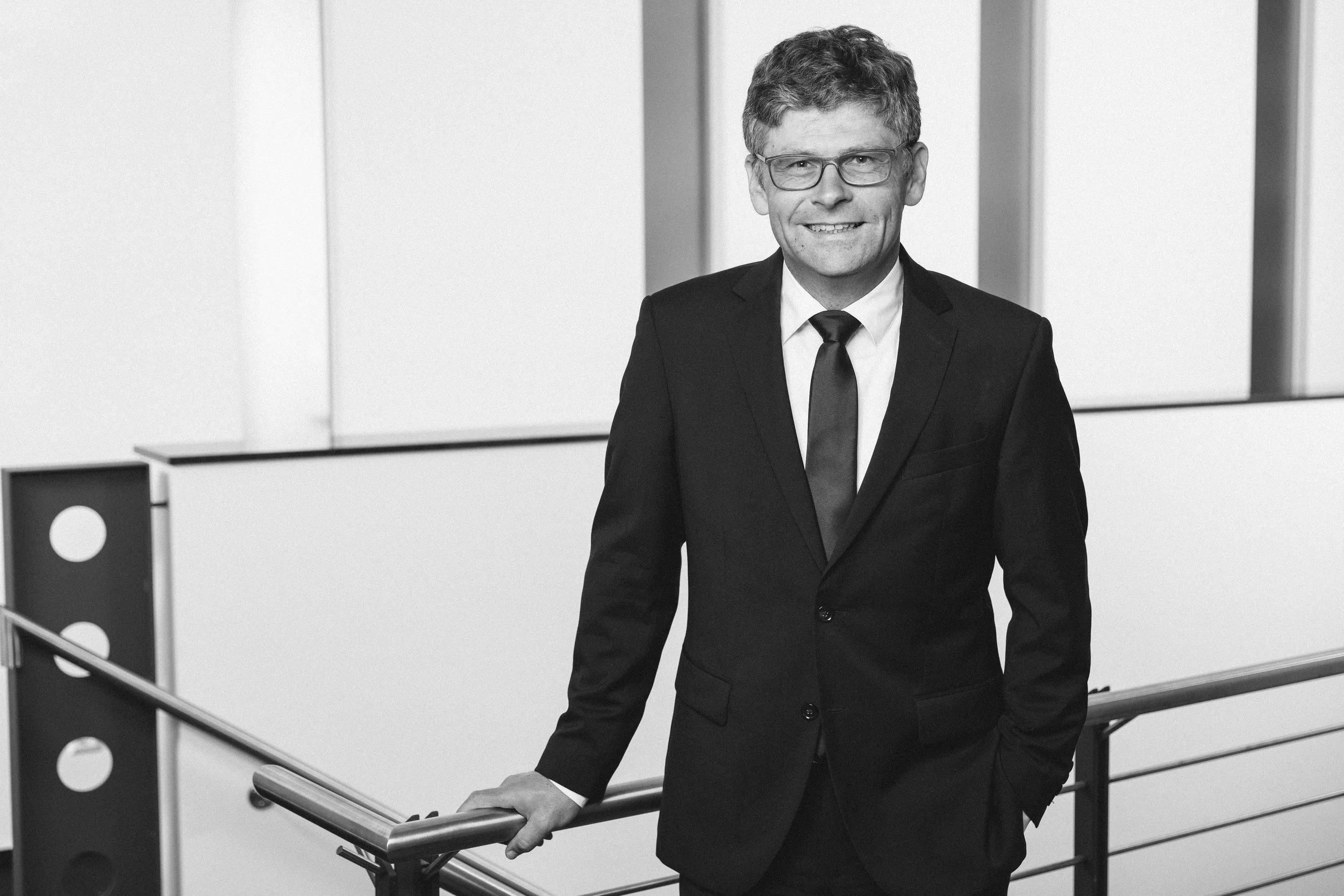 Schwarz-Weiß-Foto eines lächelnden Mannes in einem schwarzen Anzug mit Krawatte, der an einem Geländer in einem modernen Bürogebäude steht.