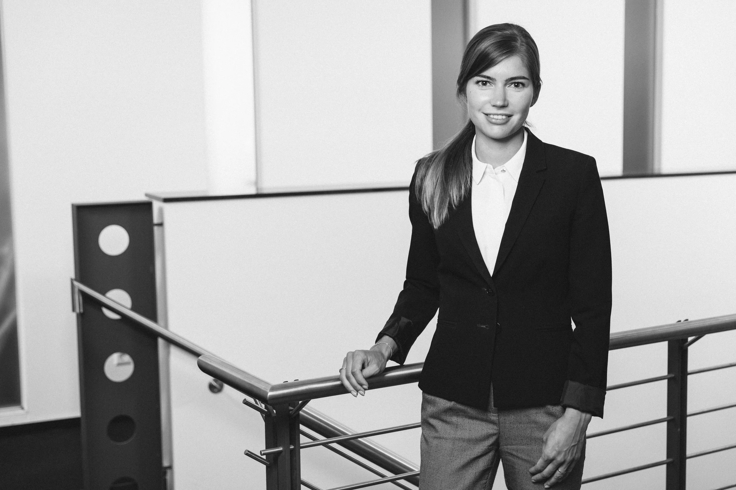 Eine junge Frau in formeller Kleidung steht an einer modernen Treppe in einem Bürogebäude, lächelt und schaut in die Kamera.