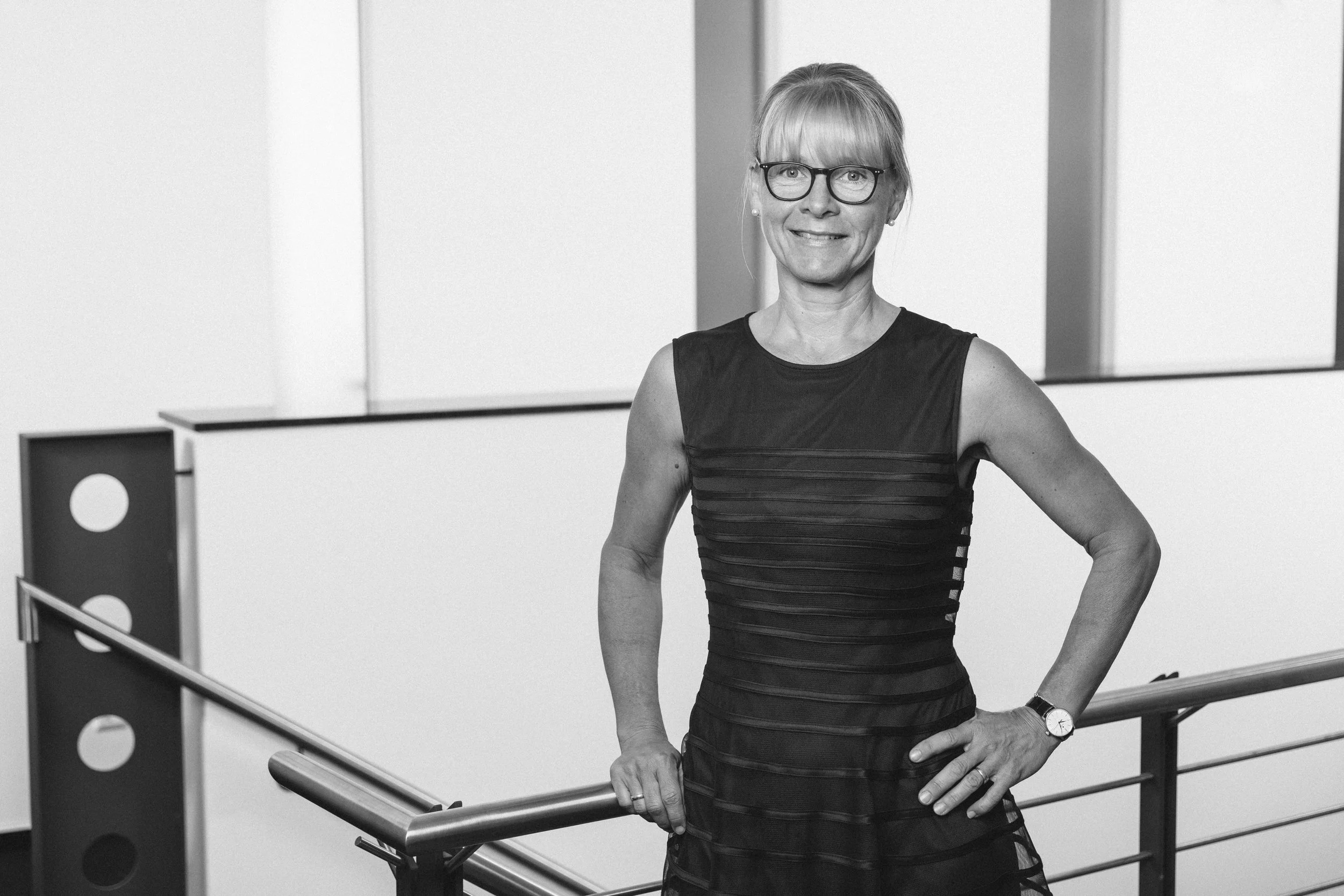 Eine lachende Frau mit Brille steht in einem modernen, hellen Gebäude. Sie trägt ein ärmelloses schwarzes Kleid und einen Uhrenarmband, hält eine Hand an die Handlauf und blickt in die Kamera.