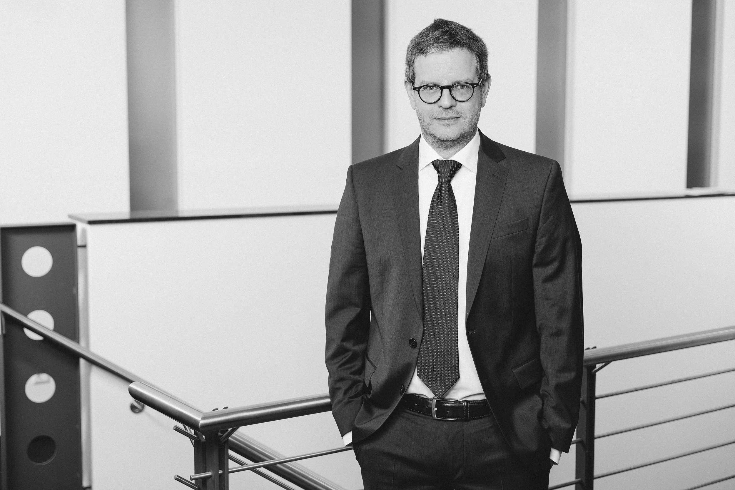 Ein Mann in formeller Kleidung, dunklem Anzug, weißem Hemd und dunkler Krawatte, mit Brille, steht an einem Geländer in einem modernen, hellen Gebäude.
