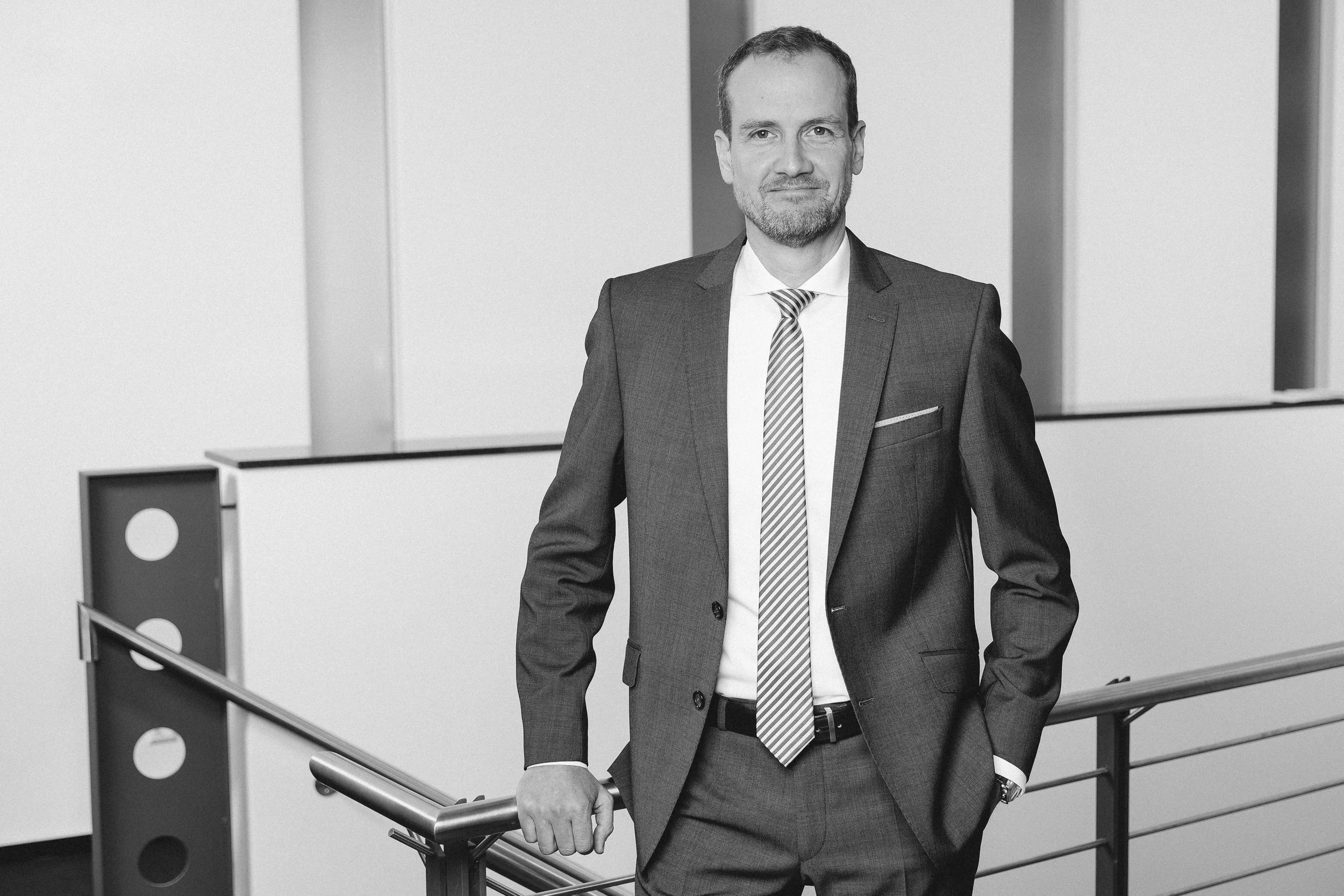 Mann in Anzug mit Krawatte steht an einem Geländer in einem modernen Büro oder Konferenzraum.