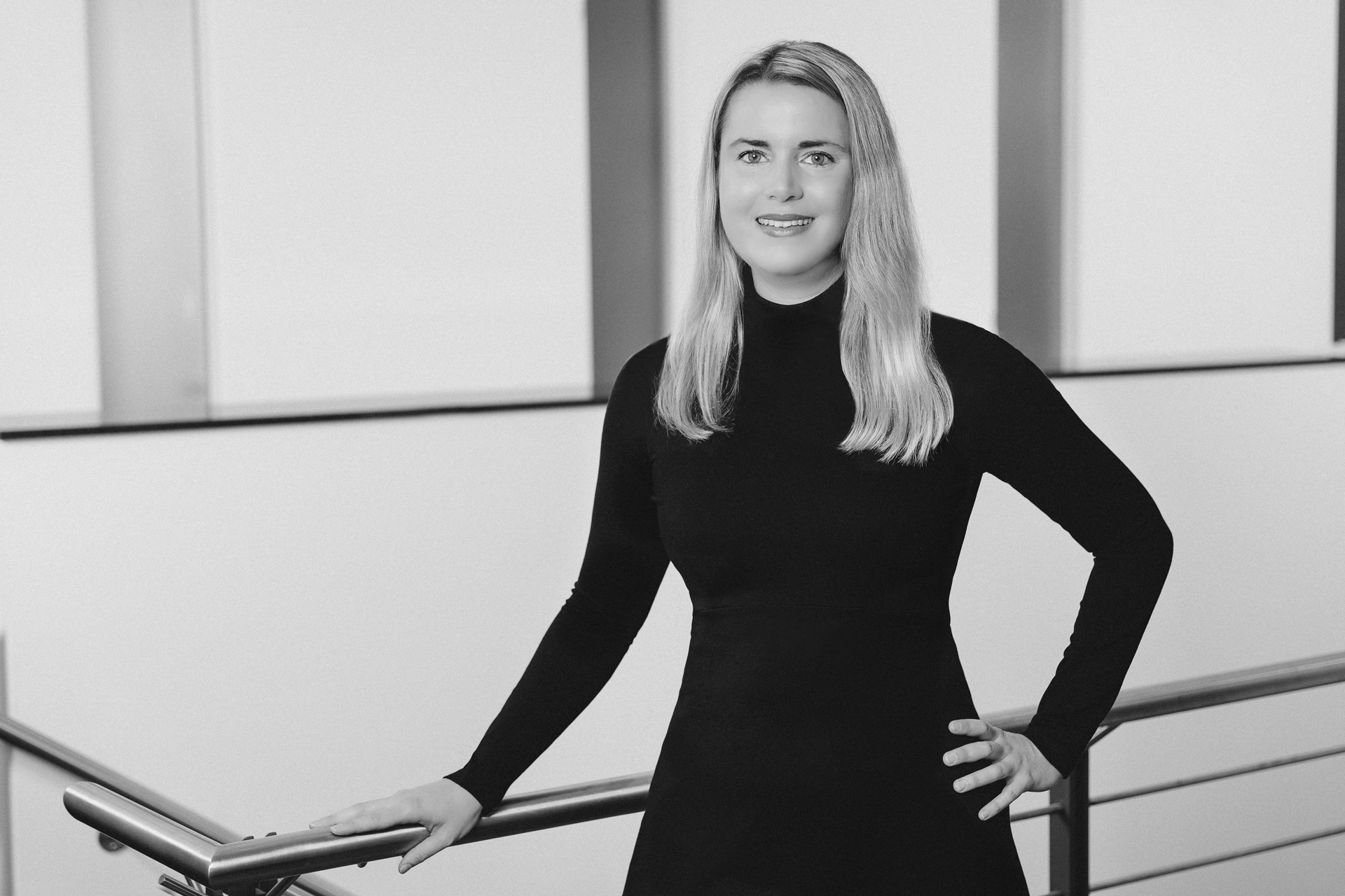 Schwarz-Weiß-Foto einer Frau mit blonden, mittellangen Haaren, die ein schwarzes, langärmeliges Kleid trägt, in einer modernen Innenarchitektur. Sie steht am Geländer einer Treppe, lächelt leicht und blickt in die Kamera.
