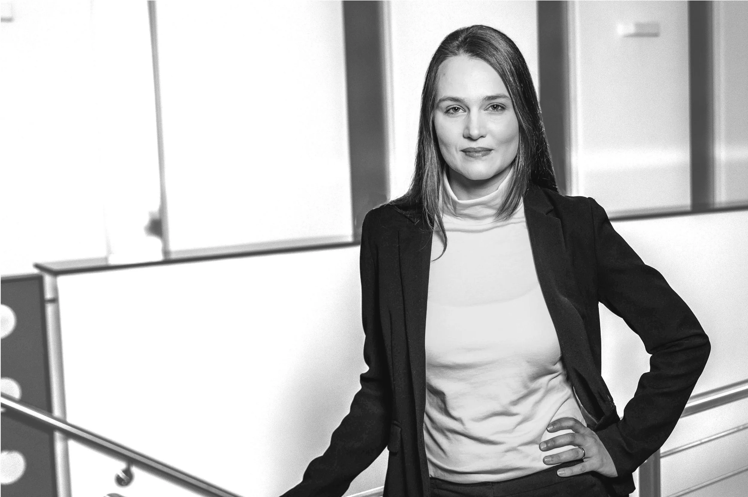 Schwarz-weiß-Foto einer Frau mit dunklem Haar, die in einem modernen Büro steht, mit einer Hand an der Hüfte, langärmliges T-Shirt und Blazer tragend.