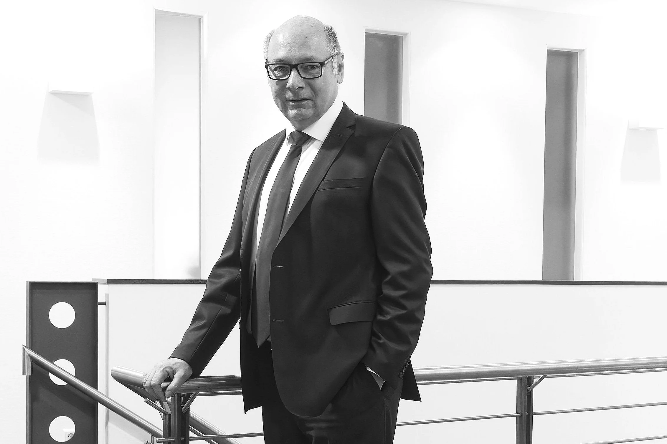 Mann in Anzug und Brille steht vor weißer Wand mit kleinen Nischen, hält sich am Handlauf fest.