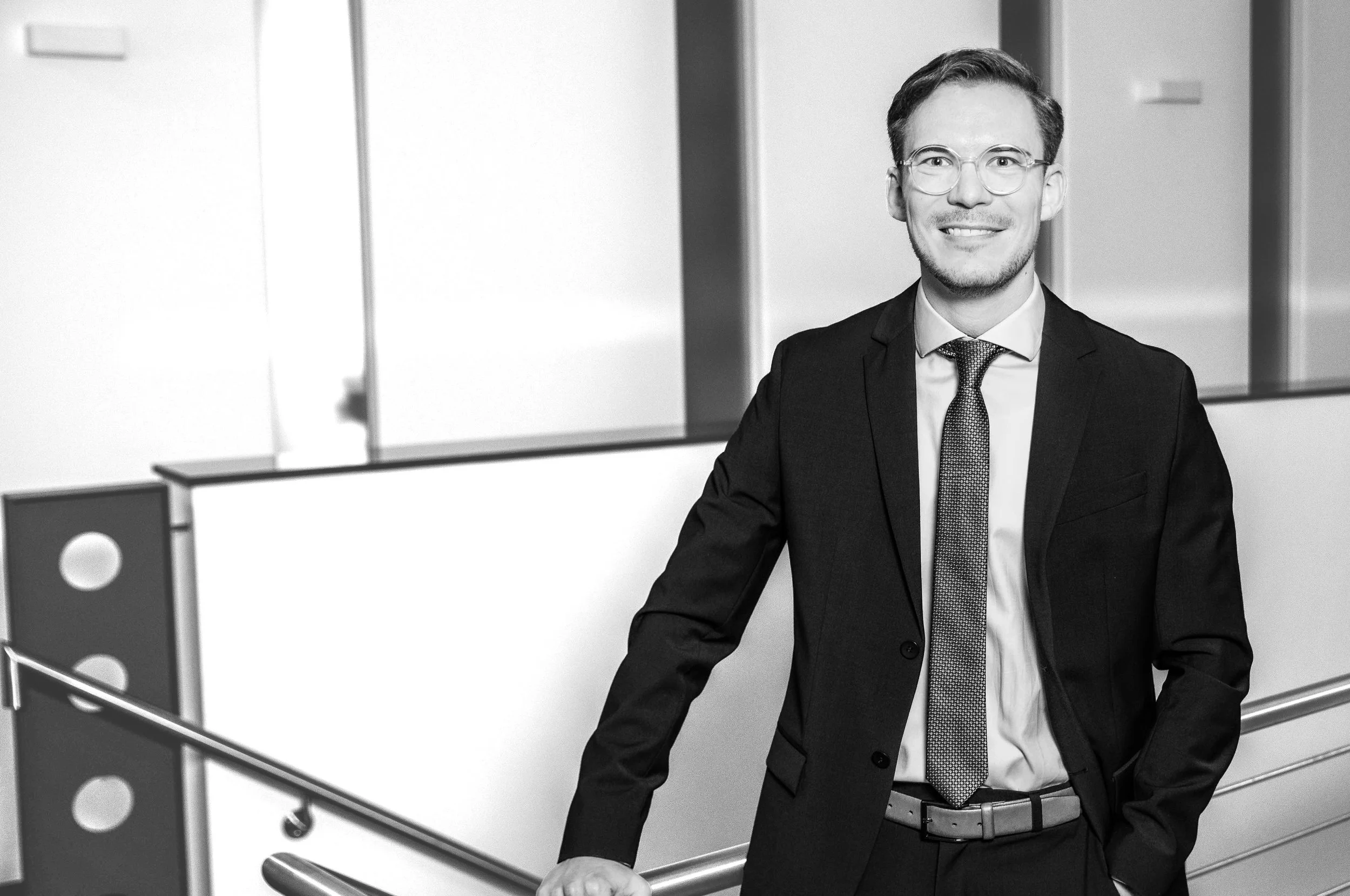 Schwarze und weiße Fotos eines Mannes in Anzug, der an einer Treppe in einem modernen Büro steht, lächelnd.