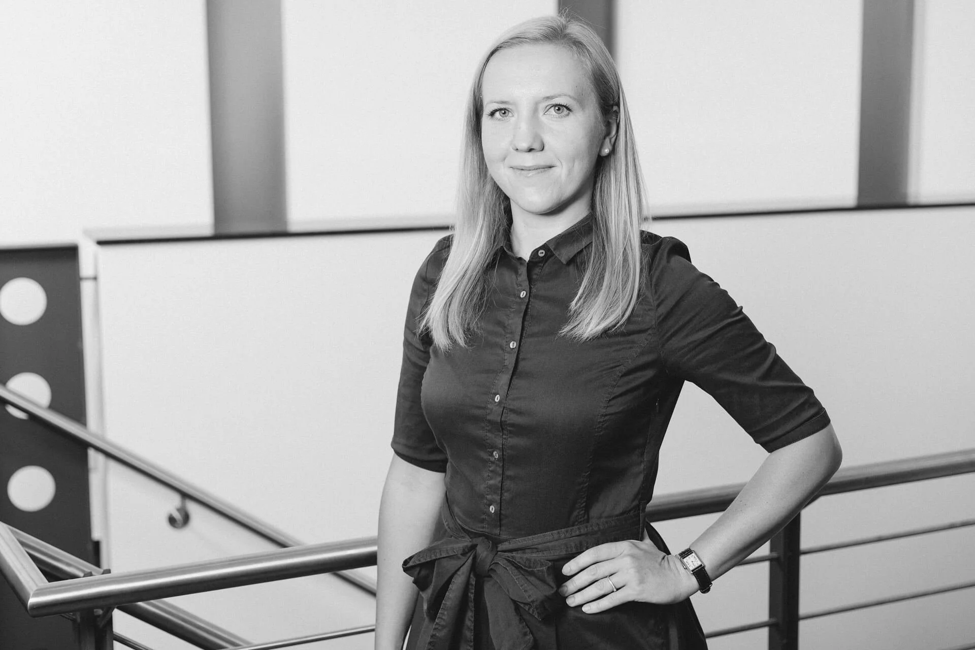 Schwarz-Weiß-Fotografie einer jungen Frau mit mittellangen, glatten Haaren, die eine dunkle Bluse trägt und an einem Handgelenk eine Uhr hat, in einem modernen Gebäude mit Metallgeländern.