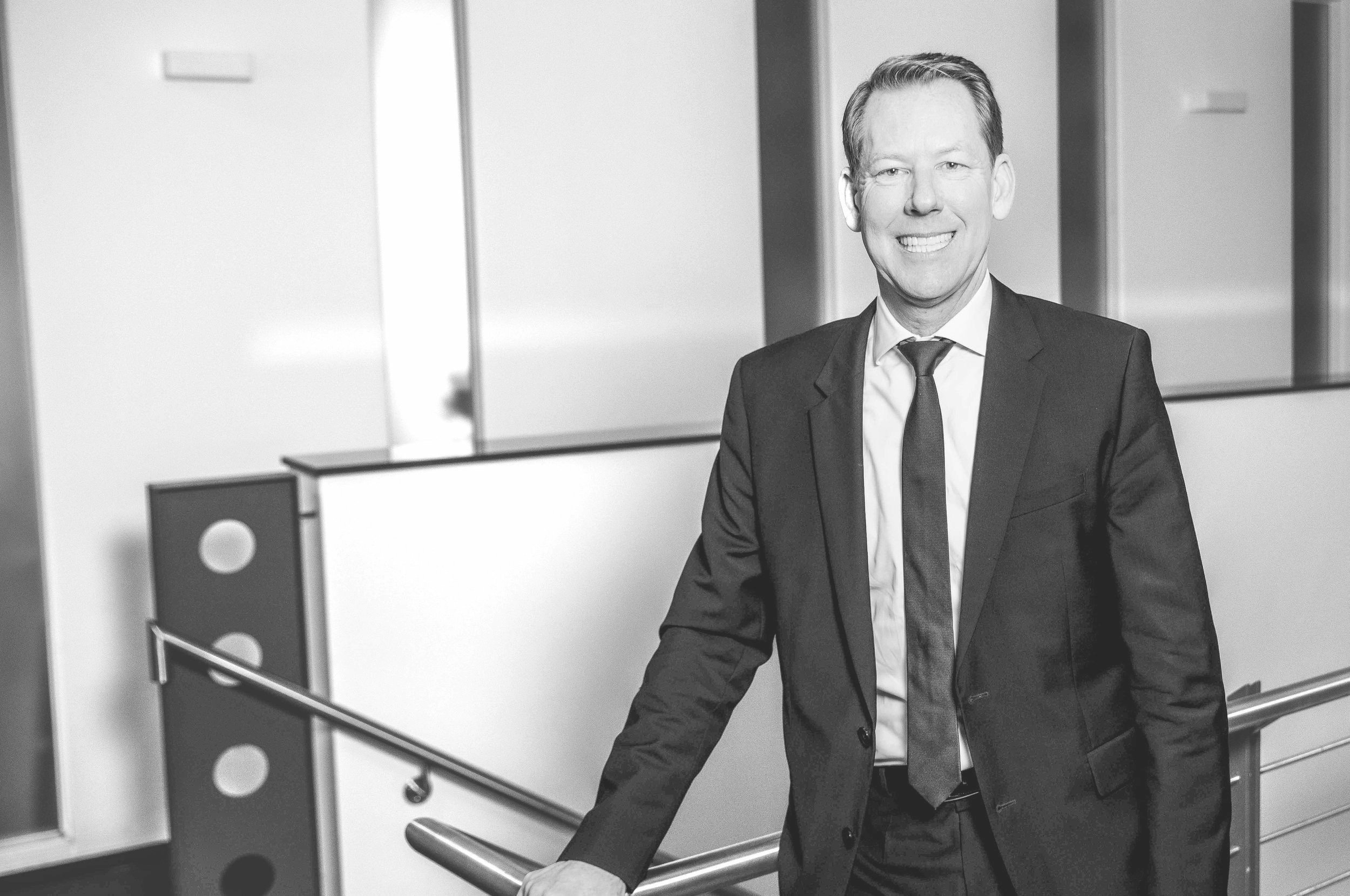 Schwarz-weiß-Foto eines lächelnden Mannes in Businesskleidung in einem modernen Bürogebäude, der die Hand auf eine Metallhandlauf stützt.