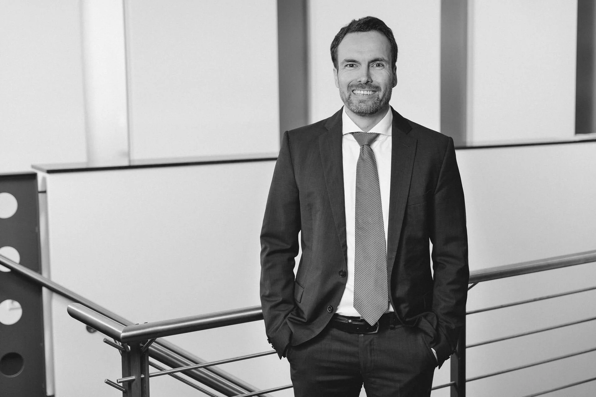 Ein Mann in einem Anzug steht vor einem Geländer in einem modernen Bürogebäude, lächelnd in die Kamera blickend.