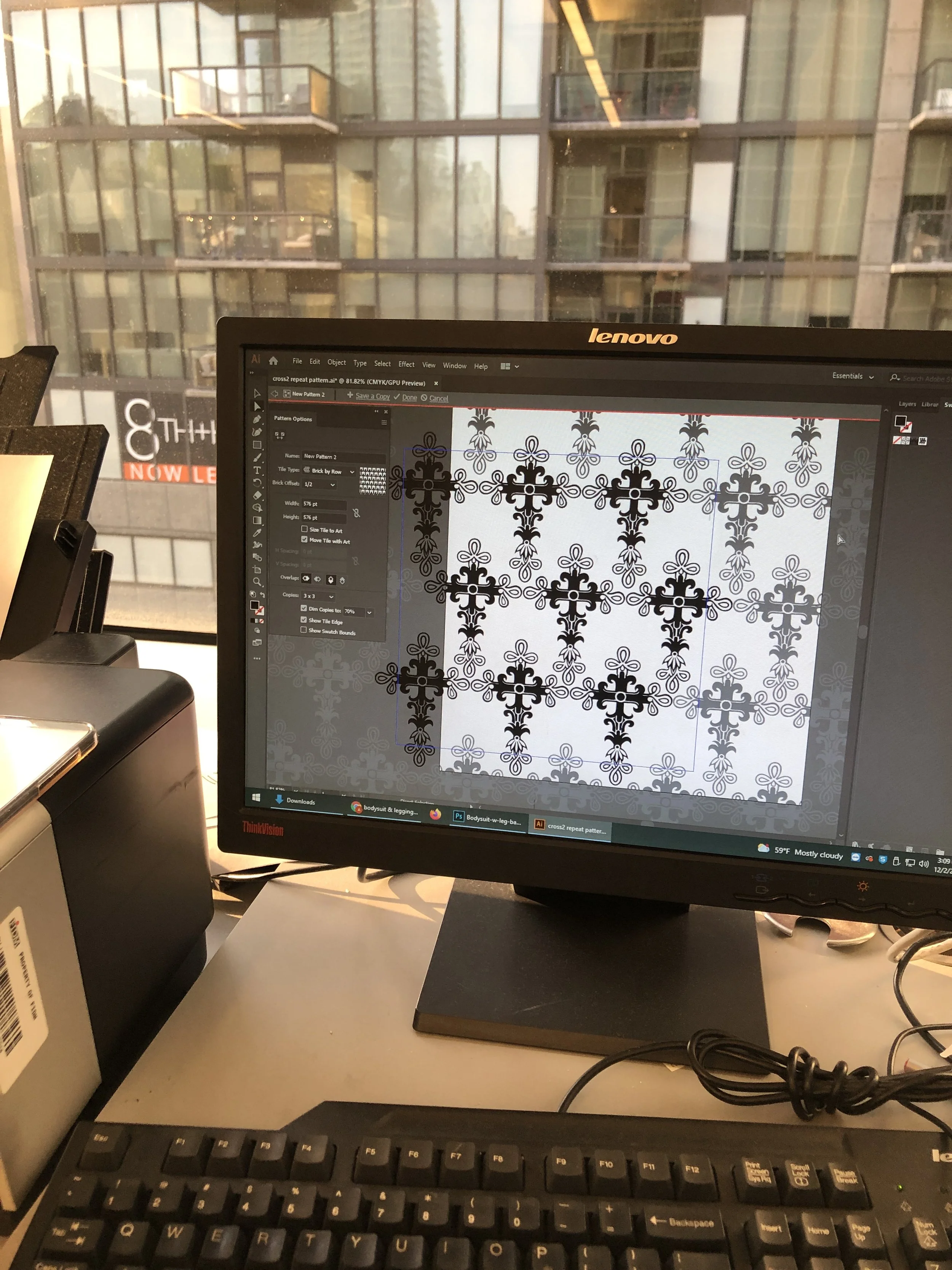 Computer monitor displaying a digital pattern with black and white intricate, floral, cross-like designs, sitting on a light-colored desk next to a black keyboard and other office equipment. A large window shows a neighboring high-rise apartment buil