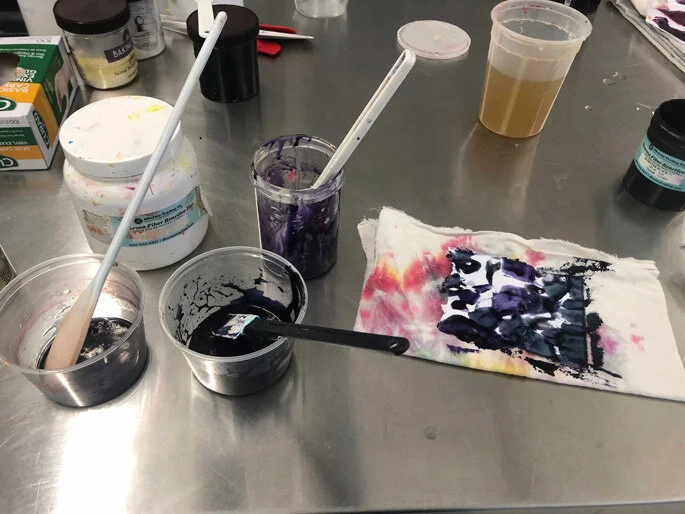 Assorted art supplies on a metal table, including small jars of black and purple paint with stirring sticks, a cloth with purple and pink paint marks, and containers of different colored liquids.