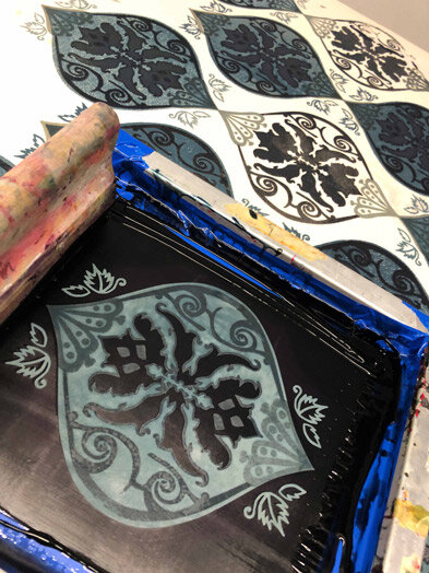 Stencil with butterfly design on black surface, with blue and yellow paint nearby, and patterned paper in the background.