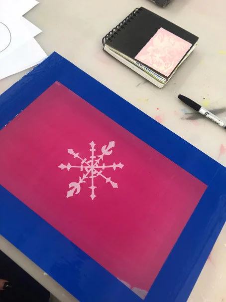 A large, pink and purple gradient sheet with a white snowflake design in the center, taped onto a blue surface. Nearby on a white table, there are notebooks, a black marker, and some paper and paint splatters.