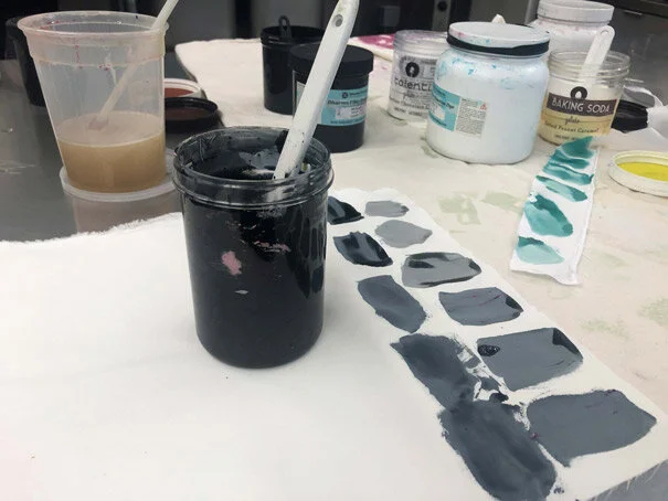 Paint palette with various shades of blue and gray, jars of paint, and a glass of water on a art table in an art studio.