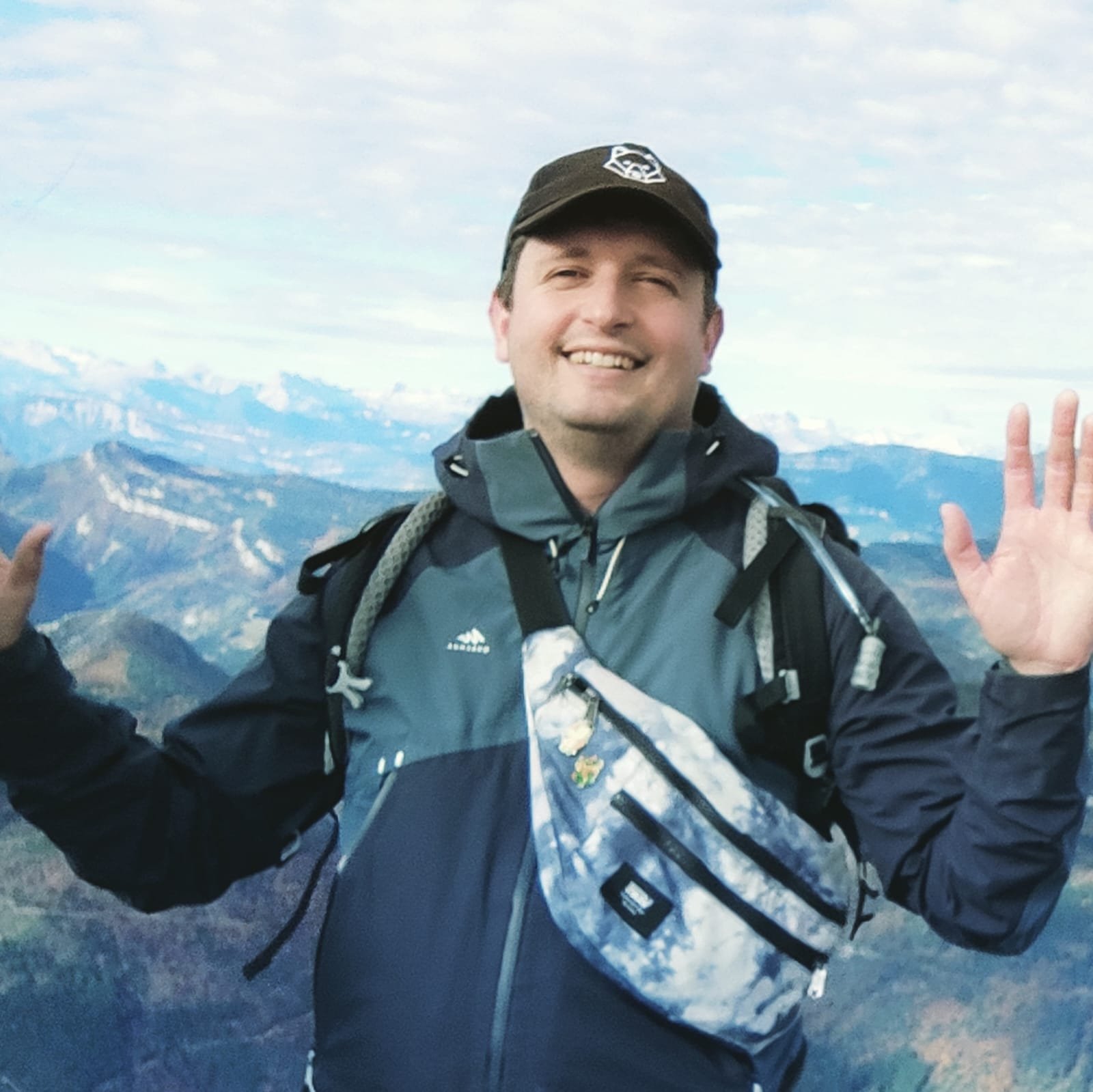 Homme souriant portant un sac à dos, faisant un geste d'accueil, avec des montagnes en arrière-plan.
