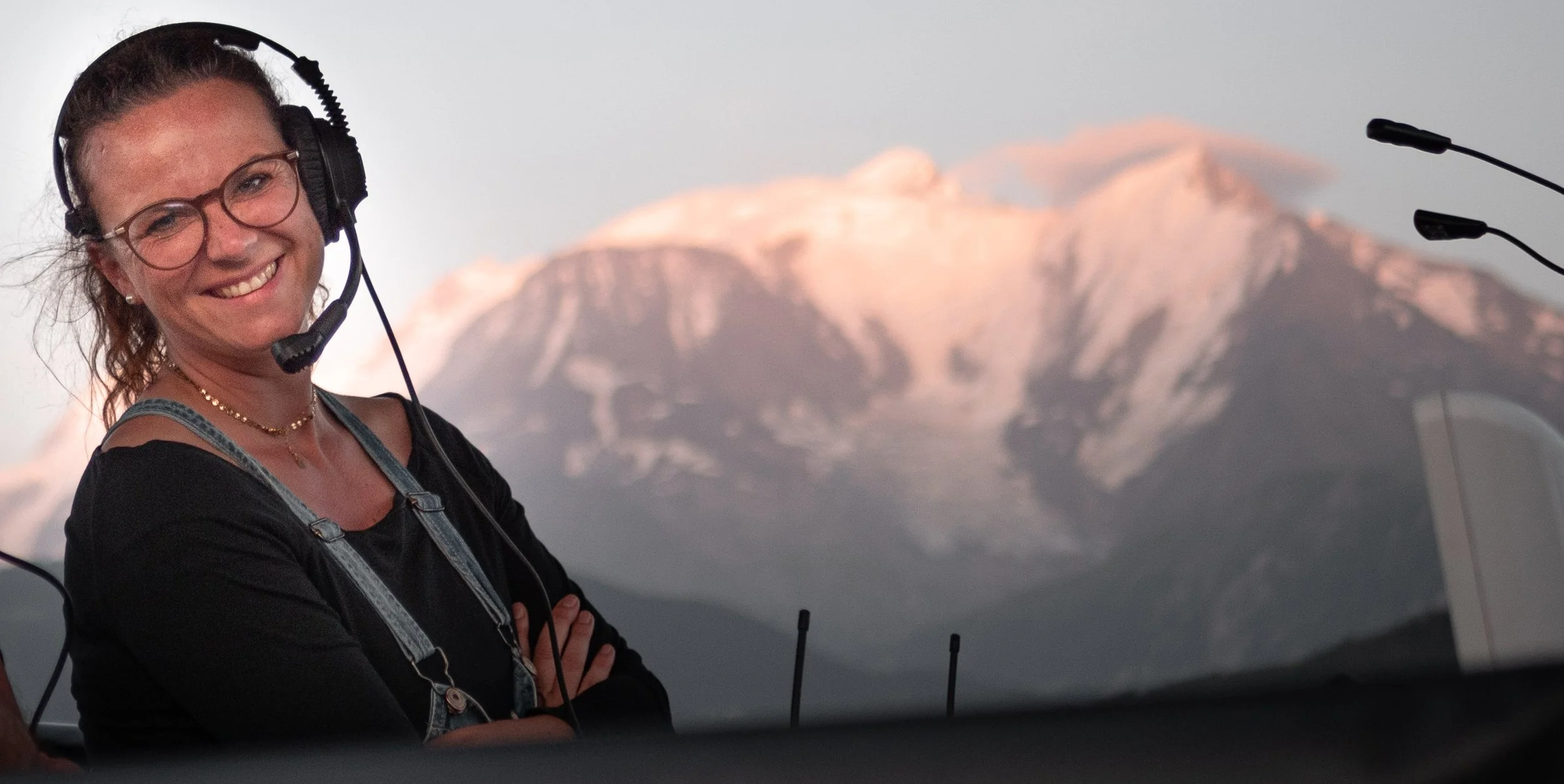 Une femme souriante portant un casque avec micro, assise devant un micro, avec des montagnes enneigées en arrière-plan.