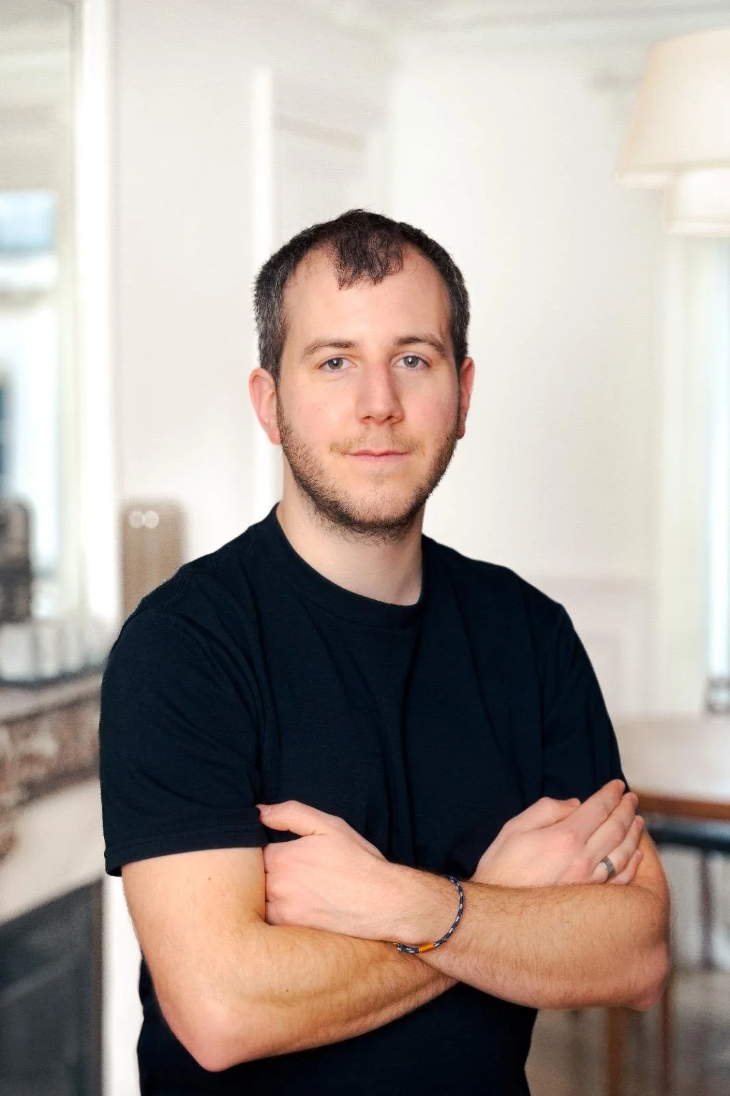 Jeune homme avec barbe, portant un t-shirt noir, croisant les bras, dans une pièce lumineuse avec des lampes et un miroir en arrière-plan.