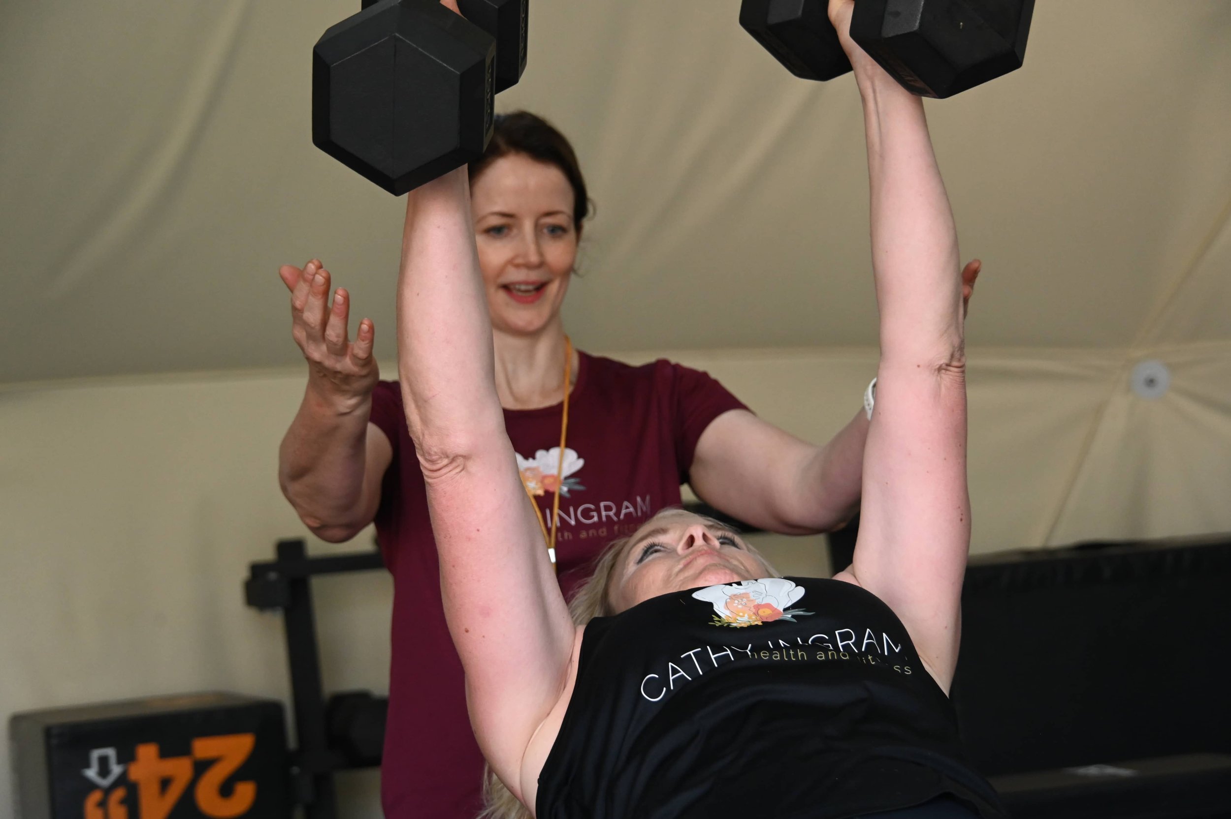 Woman assisting a person with physical therapy by holding their arms while lying on a bench with a workout sticker, in a gym or therapy center.