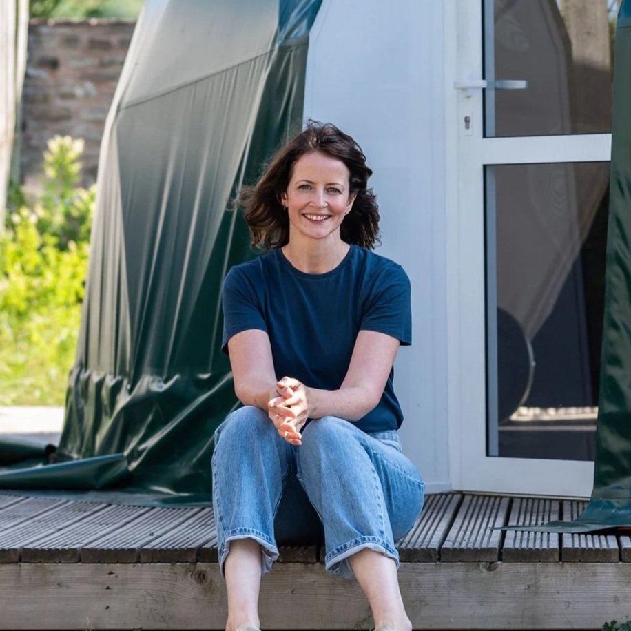 A woman with brown hair wearing a blue t-shirt and jeans sitting on a wooden deck outside a camper trailer, smiling at the camera.