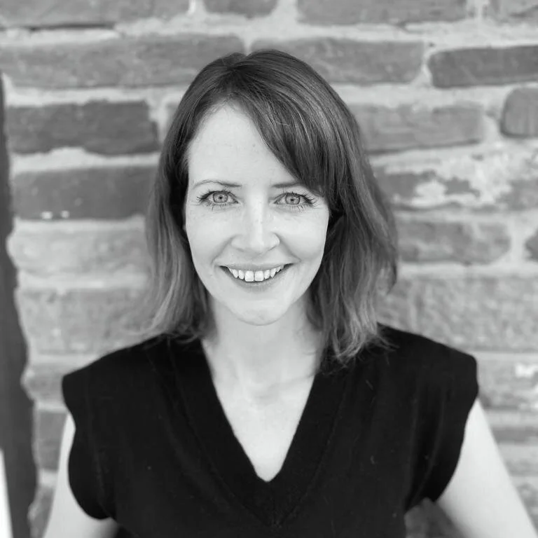 Black and white photo of a smiling woman with shoulder-length hair, wearing a sleeveless top, standing in front of a brick wall.