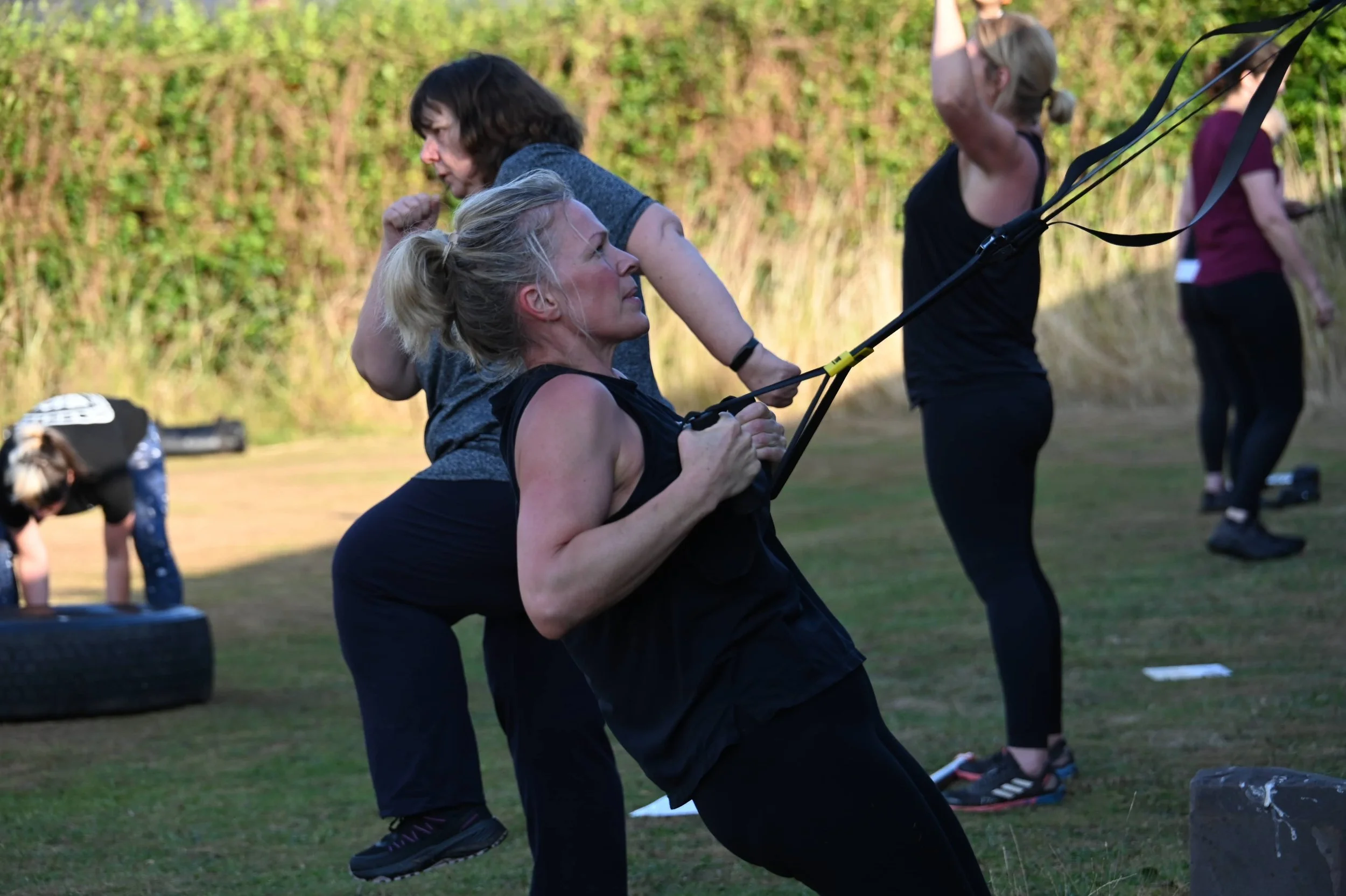 Group of women participating in an outdoor fitness class, doing squats with resistance bands, in a grassy area with trees in the background.